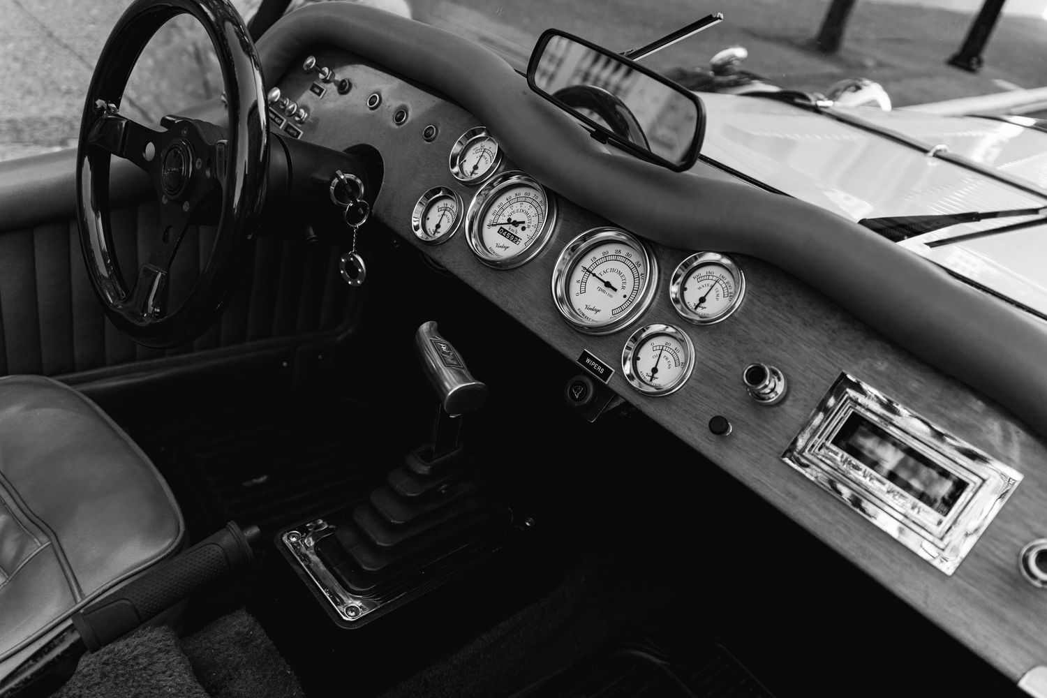 Black and white photo of a vintage car dashboard showing analog gauges and controls in a classic interior.