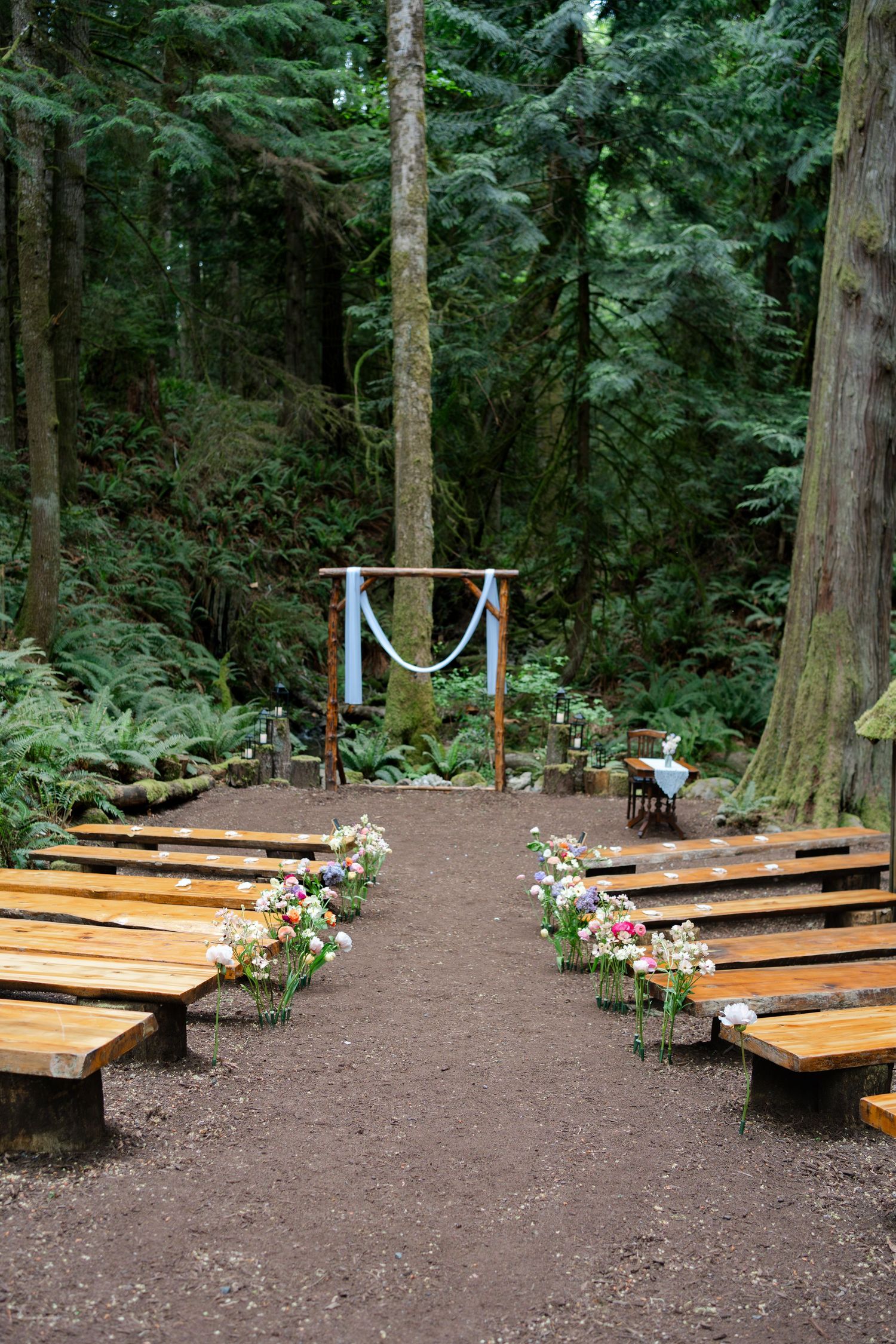 Outdoor forest wedding ceremony setup with wooden benches, floral arrangements, and a rustic arch draped in blue fabric. Sooke BC