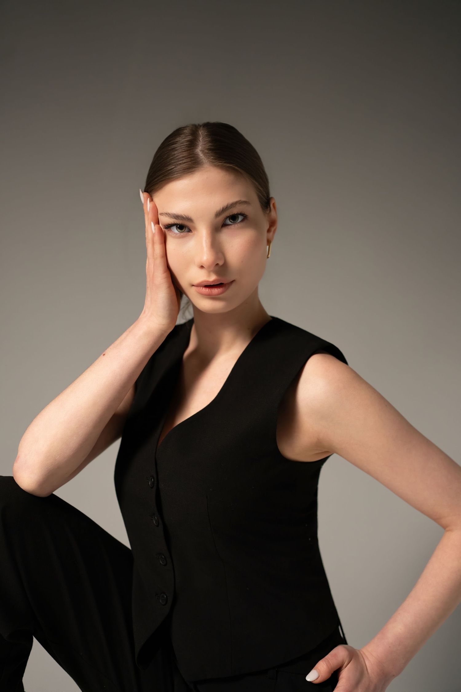 Young woman in black sleeveless vest and trousers poses confidently against gray background, hand raised to temple.