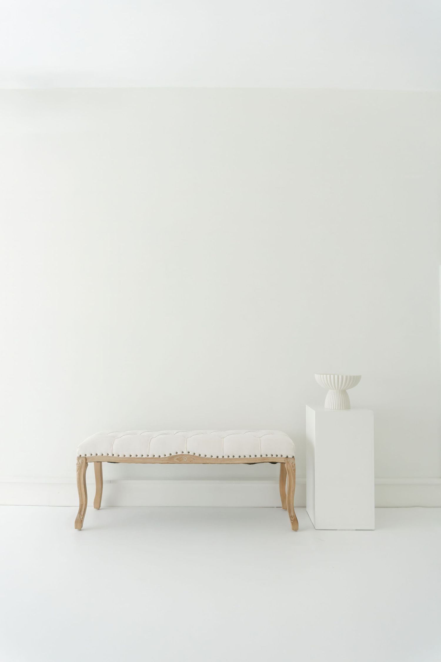 Minimalist white bench with curved wooden legs positioned against stark white wall in clean studio setting.