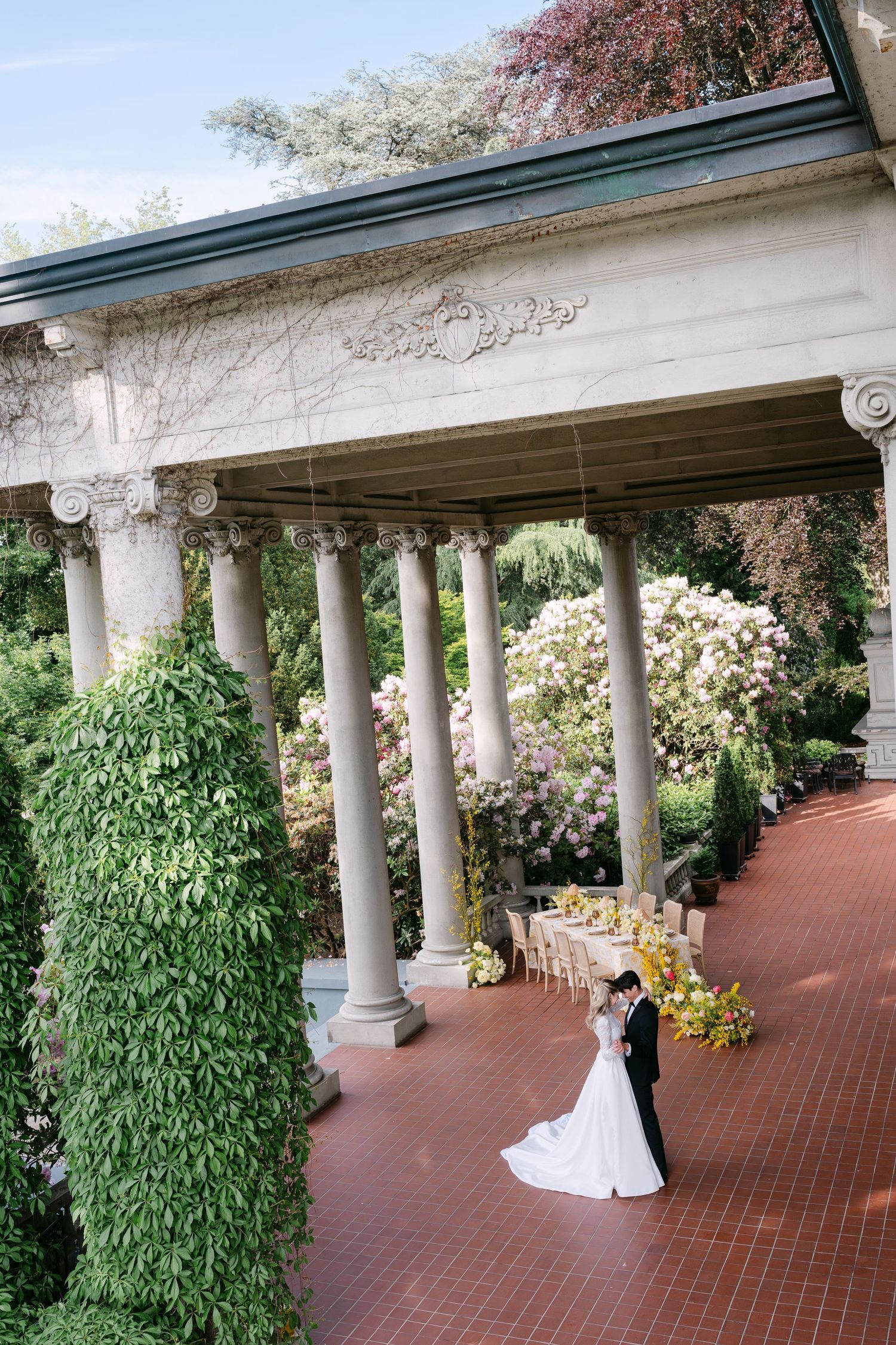 Bride and groom embrace on a brick terrace with elegant columns, lush greenery, and a floral reception table at a garden venue.