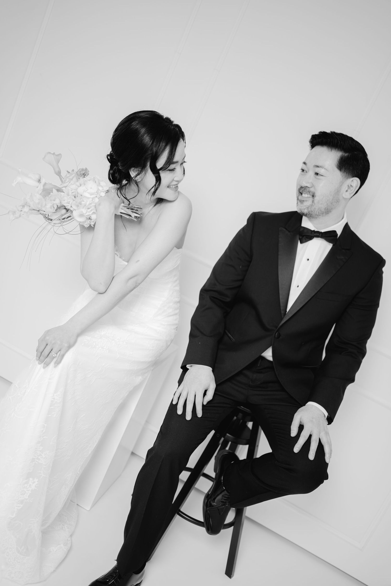 Black and white wedding photo of bride in white gown and groom in tuxedo sitting together, smiling at each other.