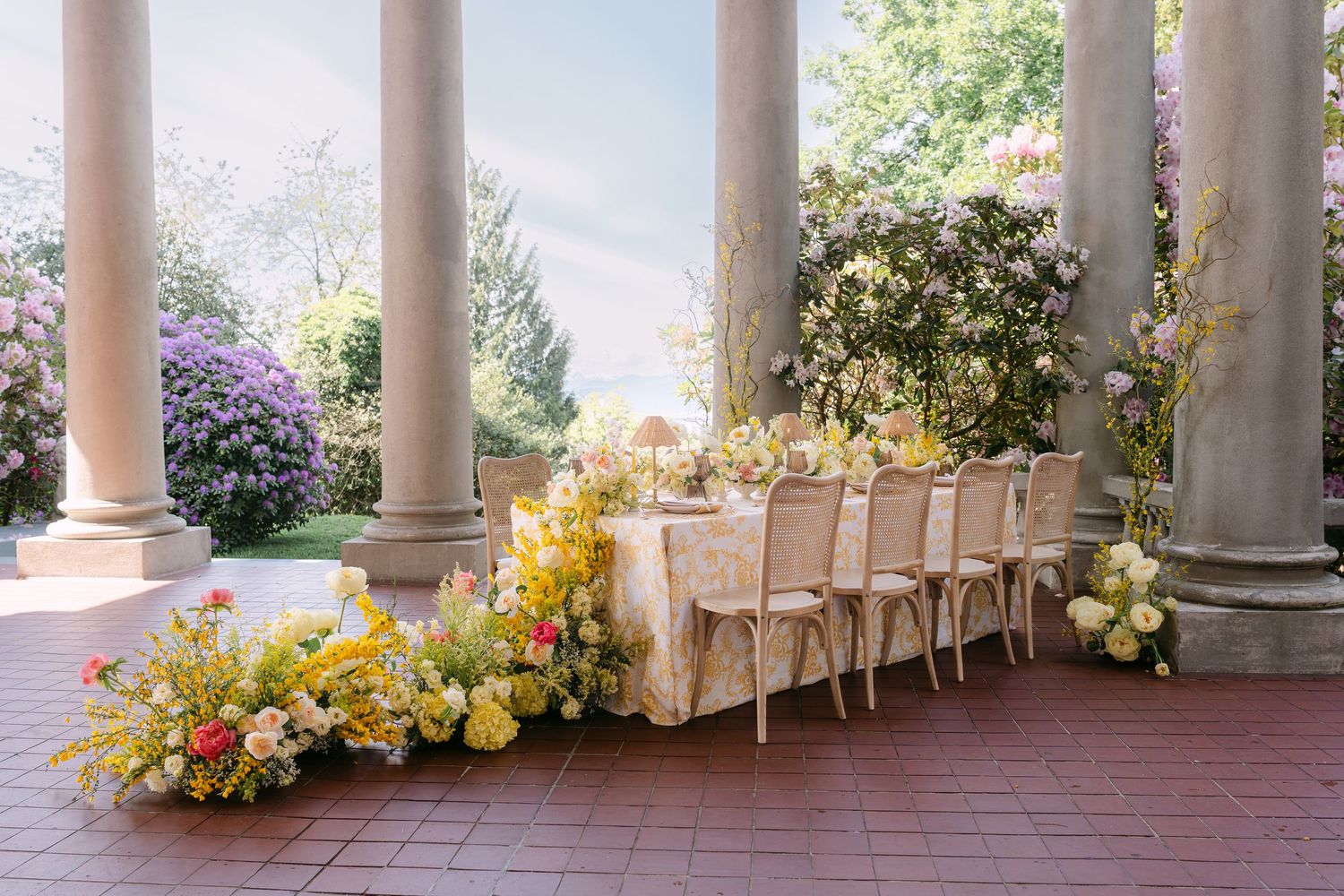 Elegant outdoor wedding reception table with yellow floral garland between grand stone columns in garden.