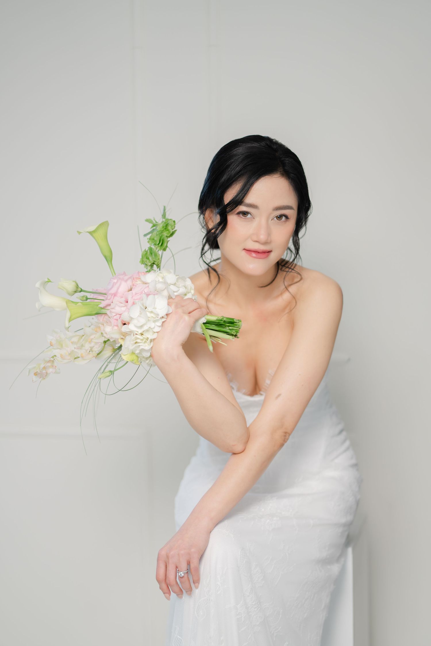 Elegant bride in white strapless gown holding a pastel floral bouquet with calla lilies and pink hydrangeas against a light background.