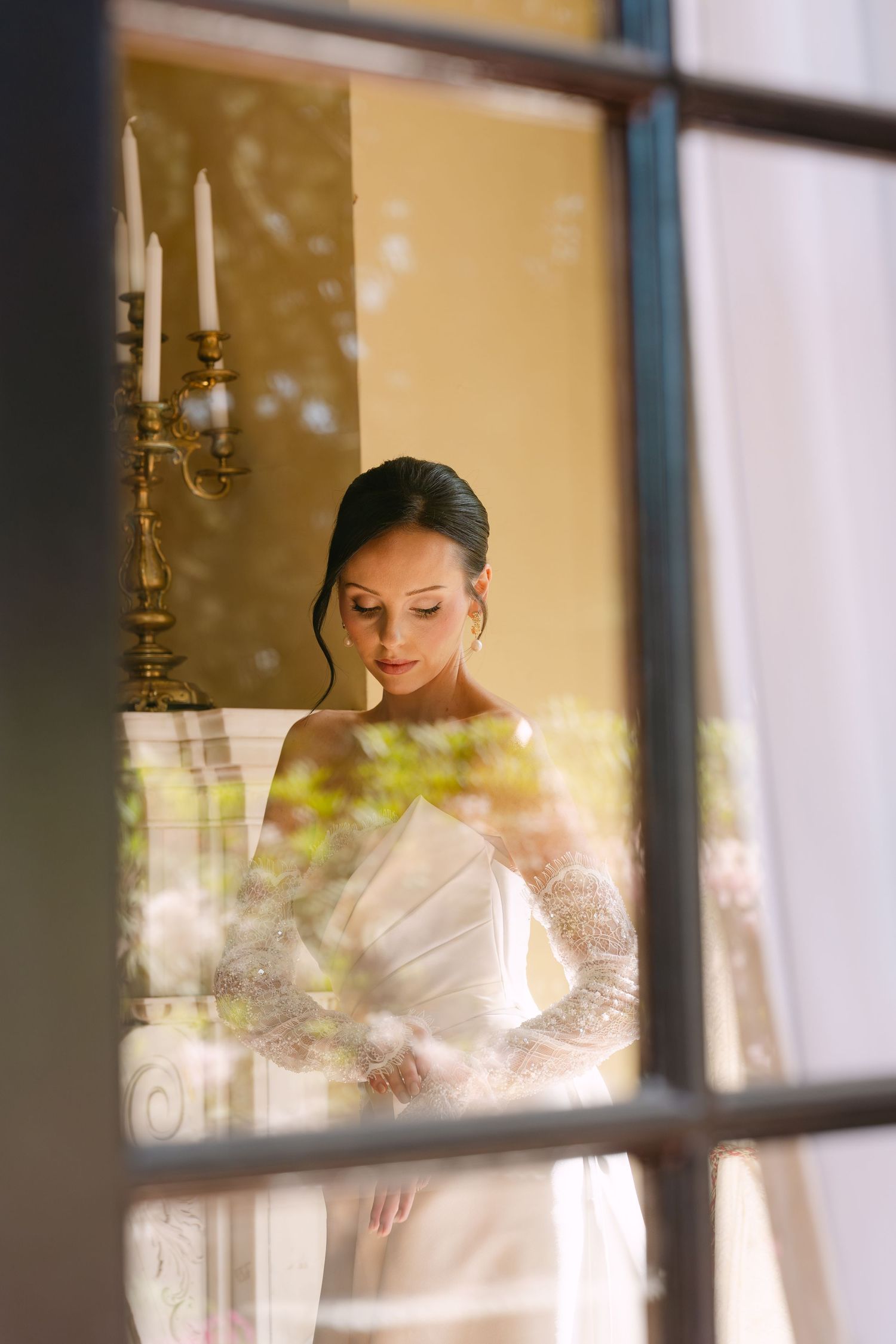 Elegant bride in white lace gown viewed through vintage window with candelabra in warm golden room.