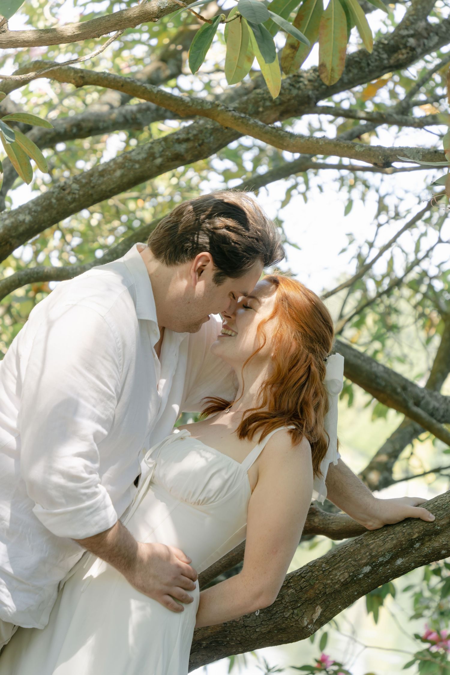 Romantic scene of two people in white clothing embracing while resting on a large tree branch.