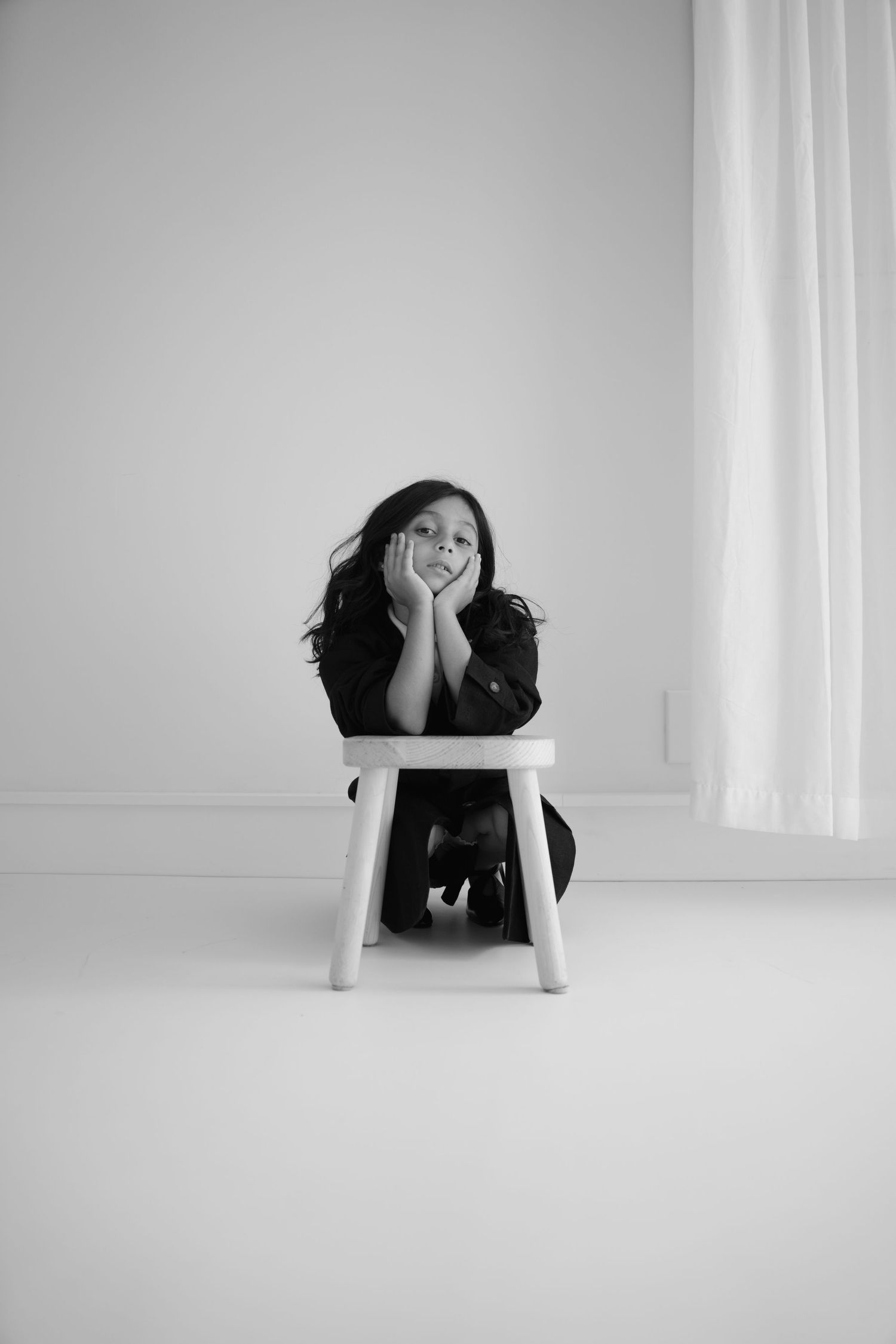Minimalist black and white portrait on small white stool against plain background.