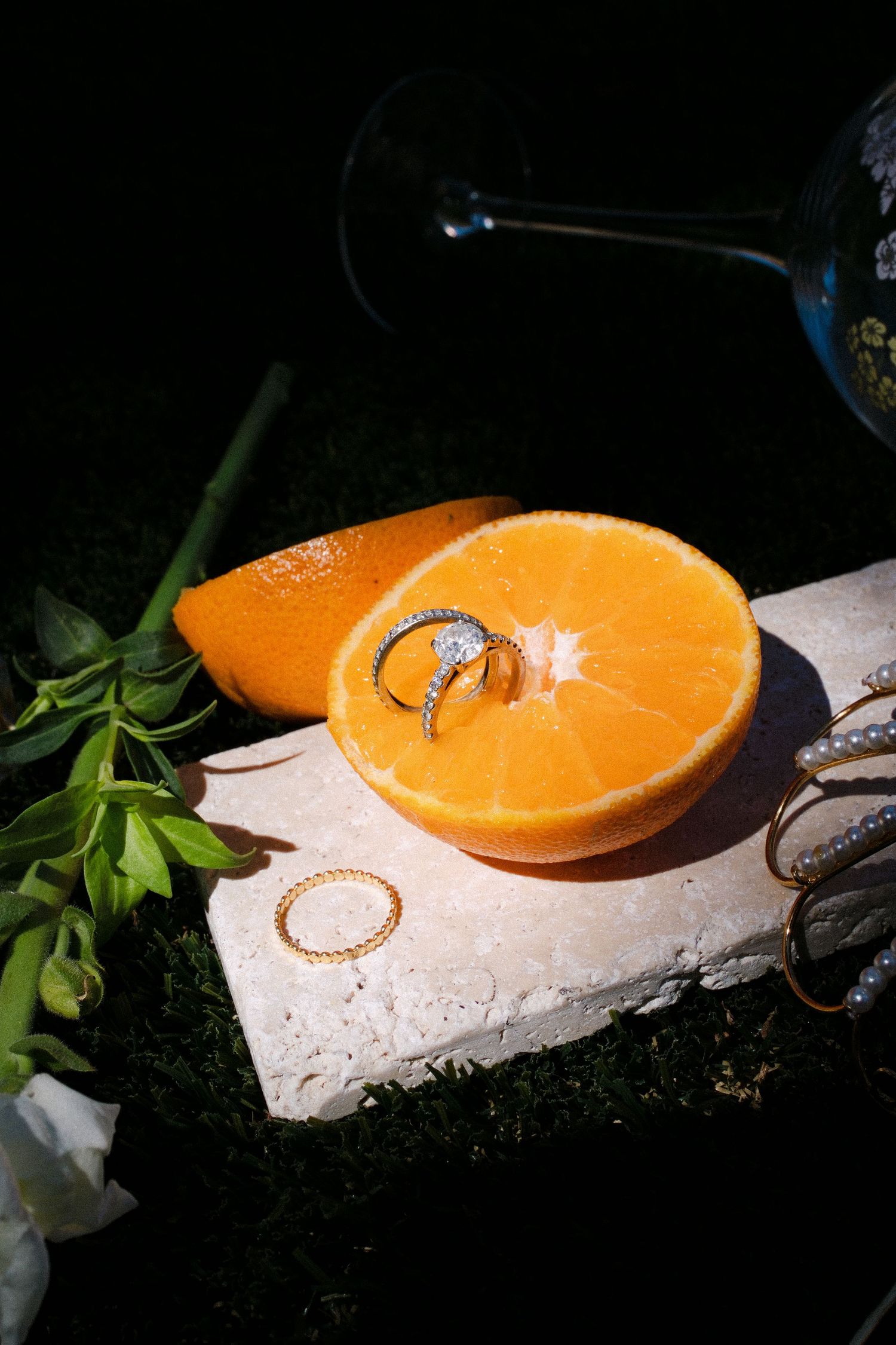 Engagement rings displayed on a halved orange atop a stone slab, with white flowers and a wine glass in the background.