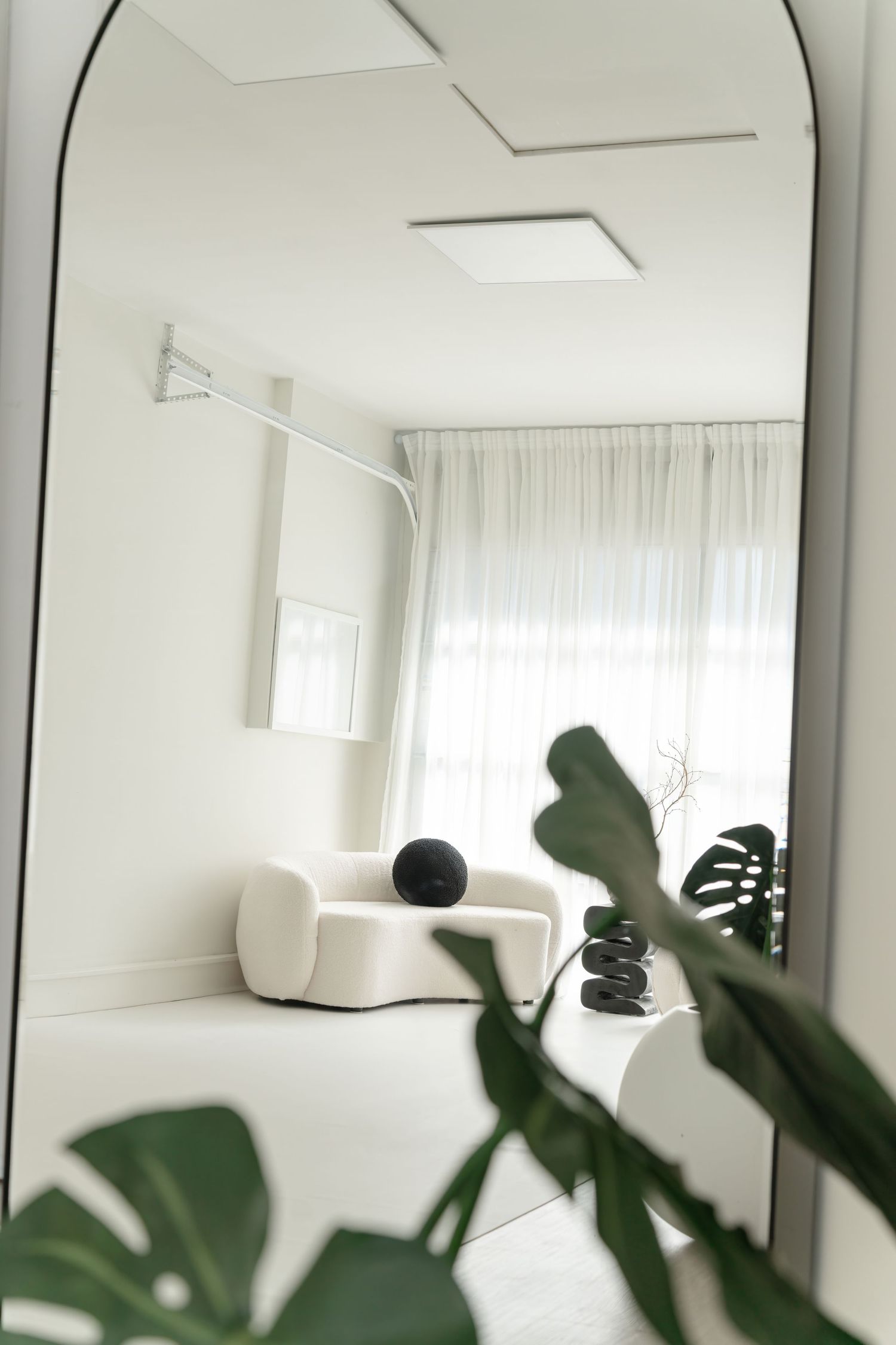 Arched mirror reflection of minimalist white room with curtains and tropical plants.