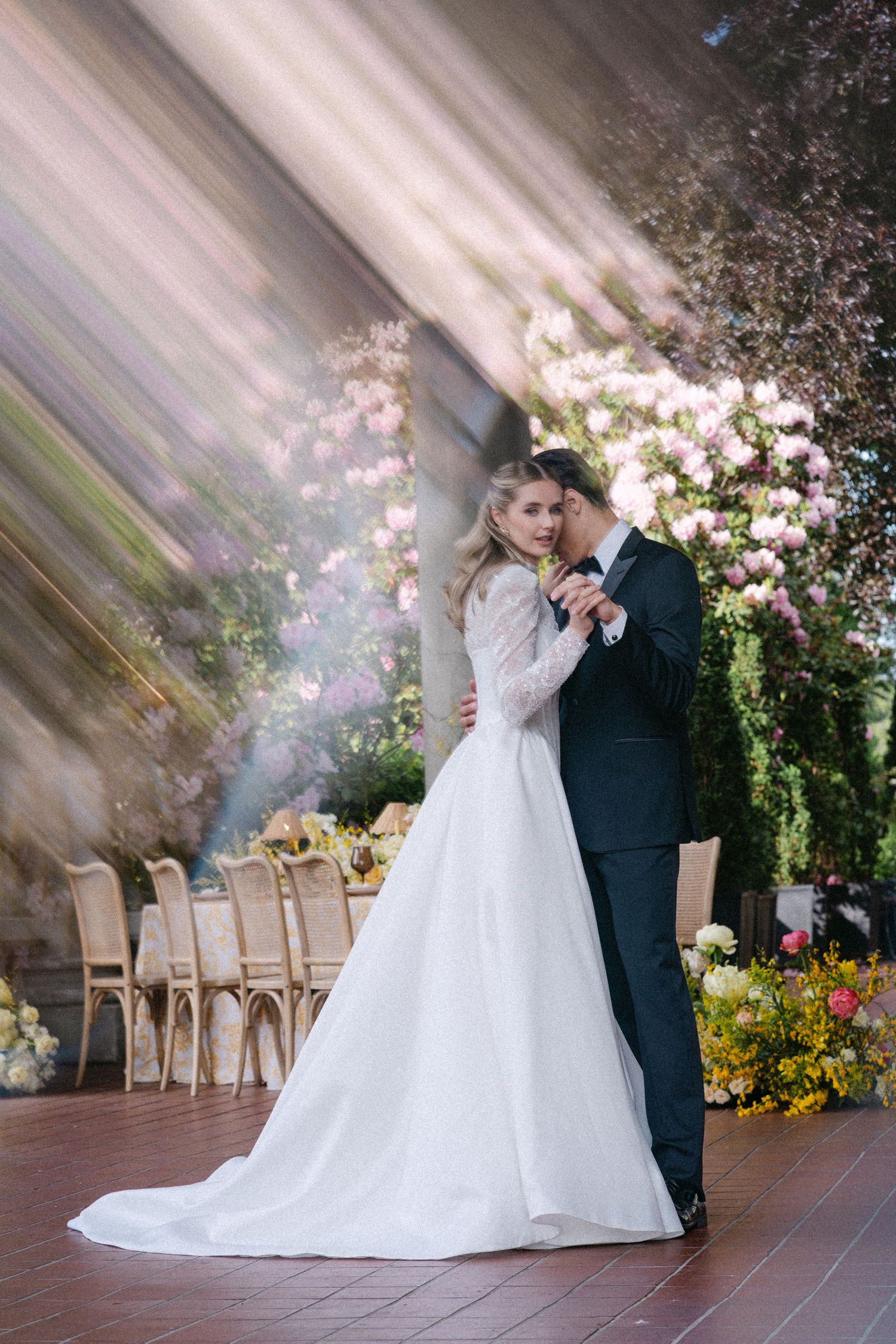 Bride and groom share first dance in dreamy venue with lush floral backdrop and soft dramatic lighting.