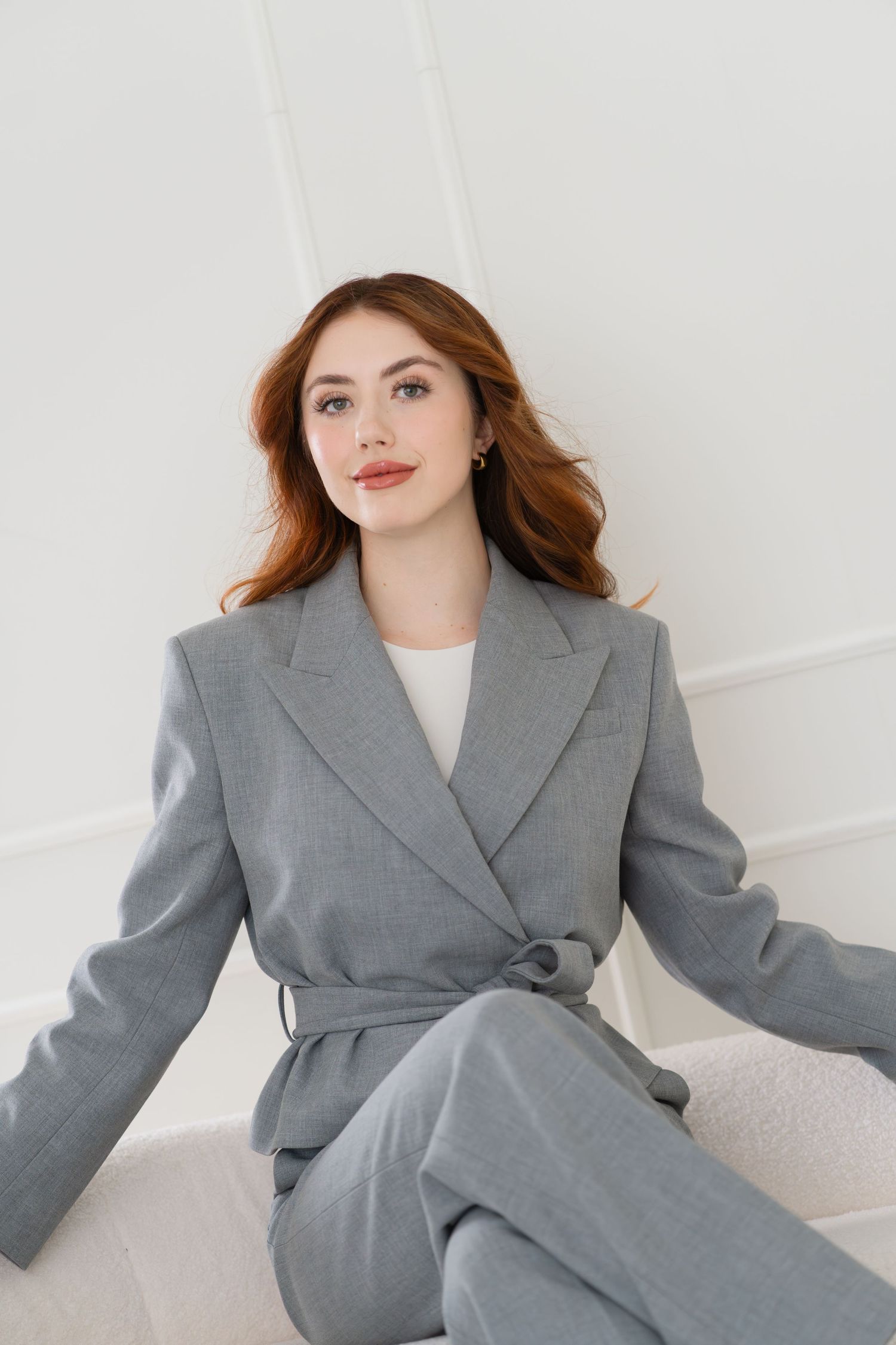Woman in gray belted blazer suit with red hair poses elegantly against a white wall.