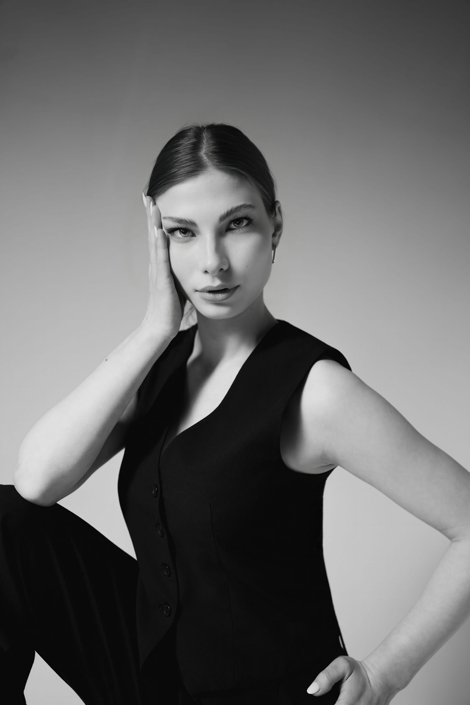 Black and white portrait of a young woman in a sleeveless black vest, posing with hand near face against gray background.