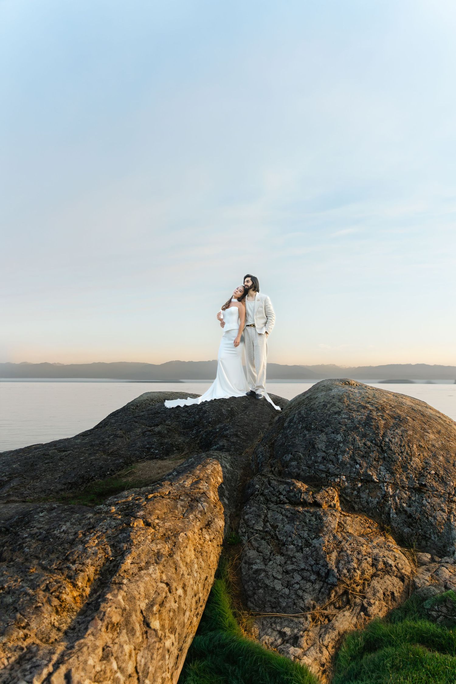 Bride and groom in white standing on rocky coastal cliffs at sunset with calm water and mountains in background.