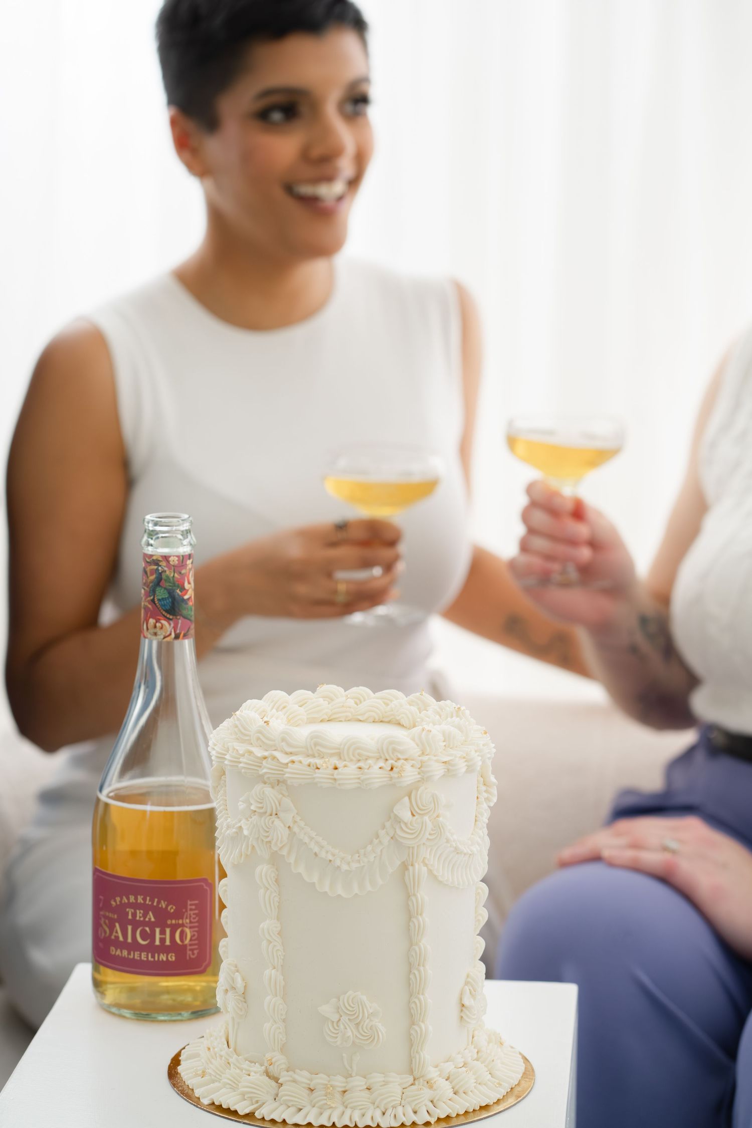 White decorated celebration cake with sparkling wine bottle, two women toasting with champagne glasses in background.