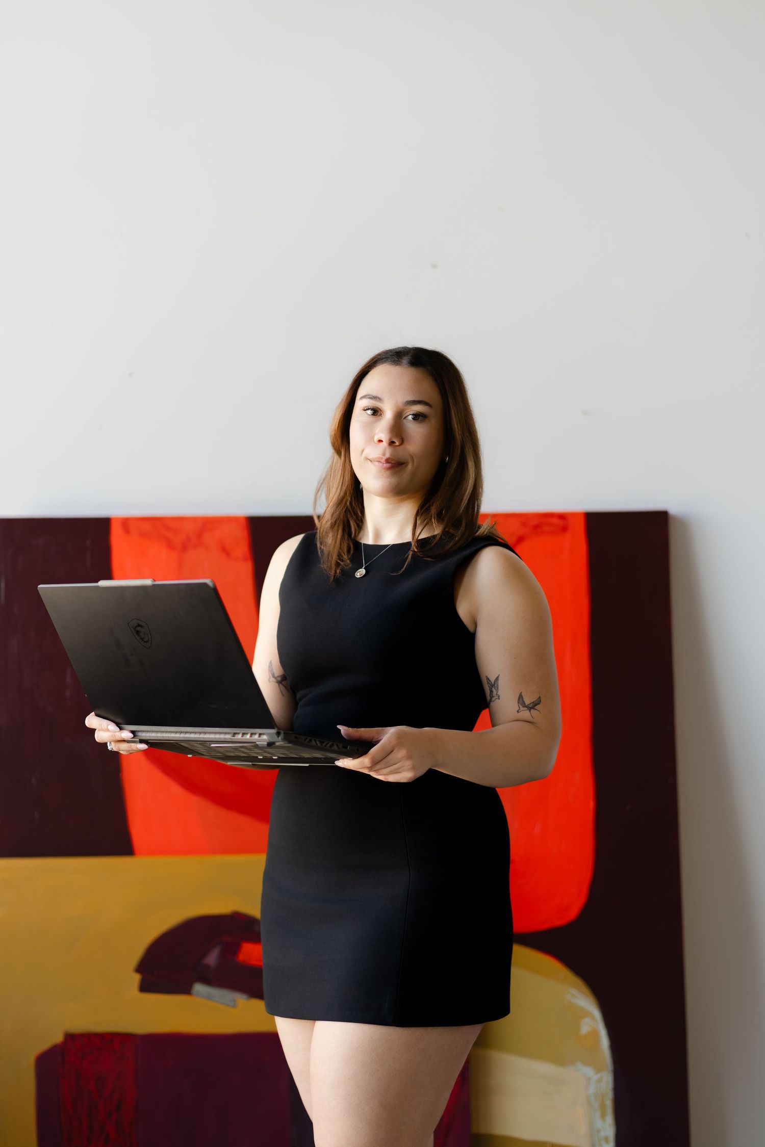Woman in black dress holding laptop stands before colorful abstract painting in bright studio.