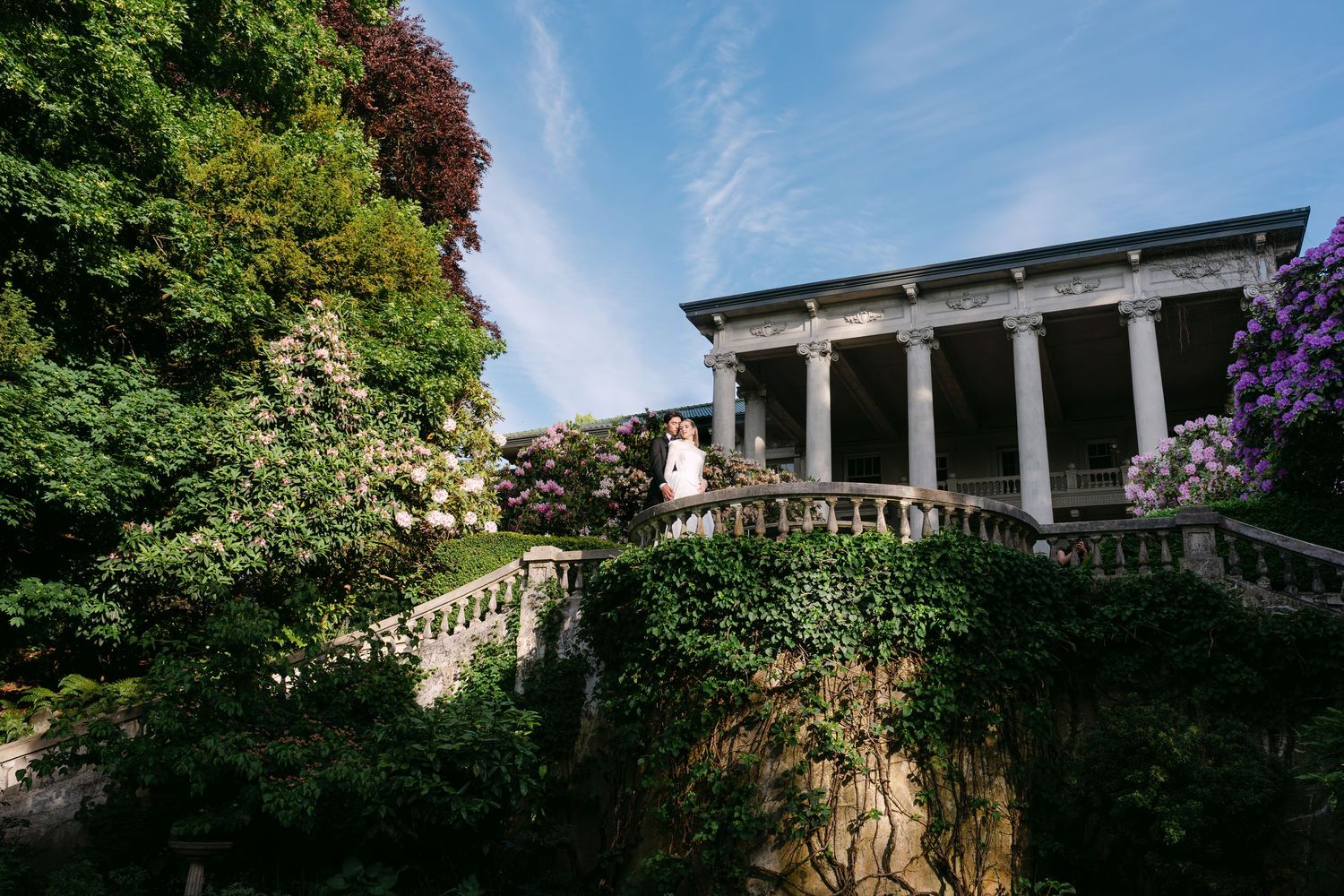 Couple poses on stone staircase of elegant villa with columns, surrounded by lush greenery and wisteria under blue sky.