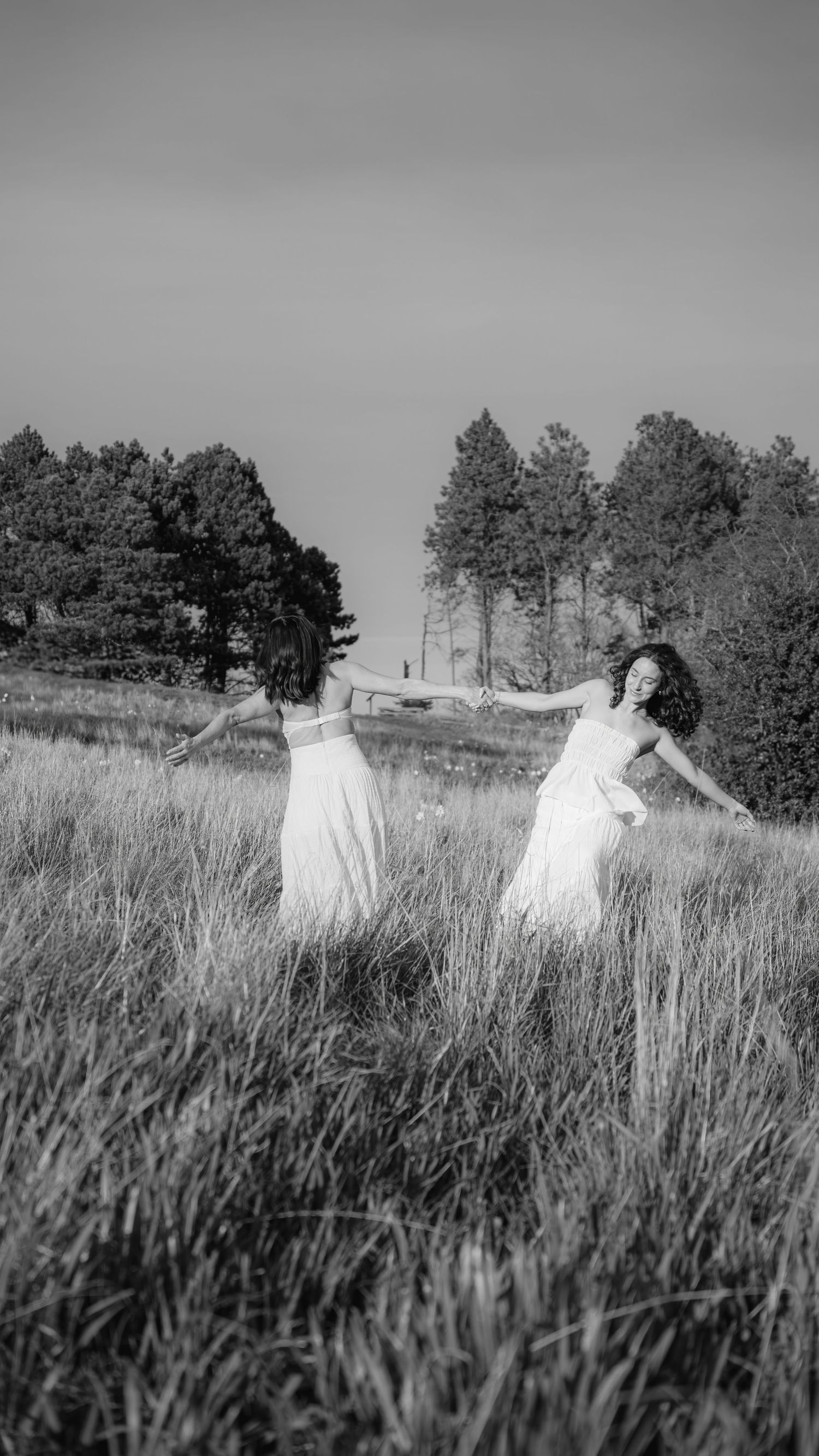 Black and white photo of two people in white dresses dancing in a meadow with pine trees.