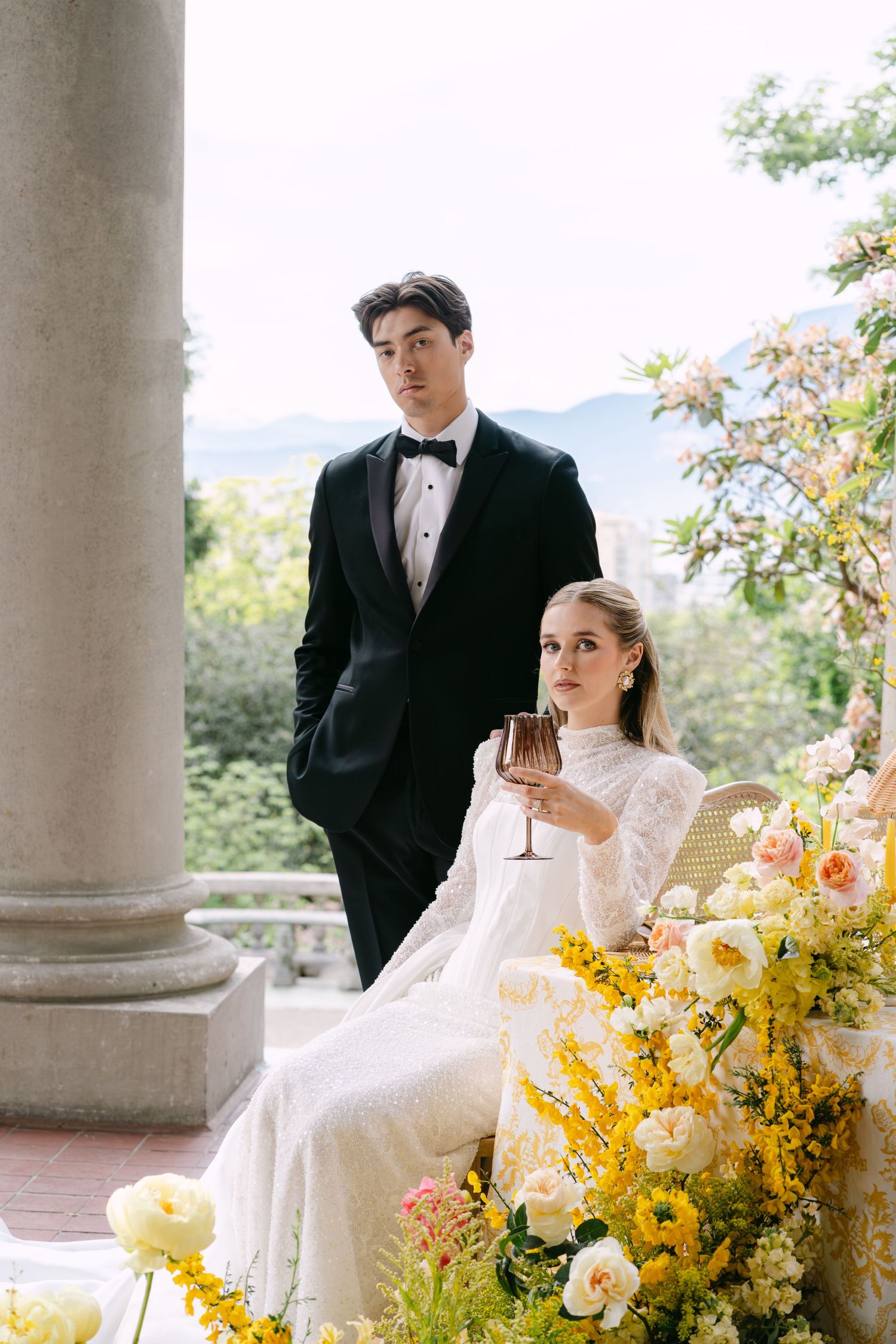 Elegant bride in lace gown holds champagne beside groom in tuxedo on floral-adorned terrace with mountain views.
