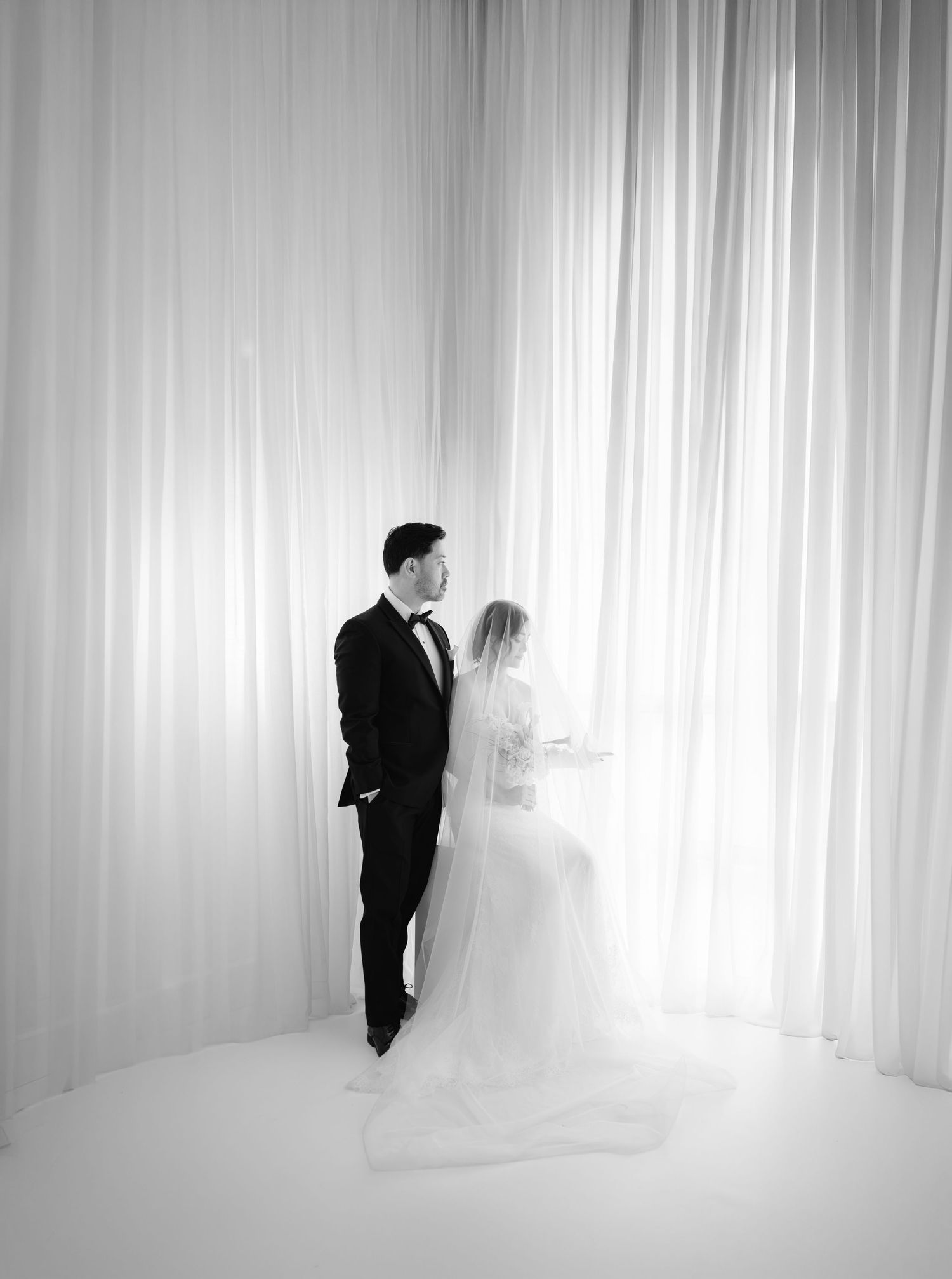 Black and white photo of bride and groom standing before draped white curtains in elegant wedding attire.