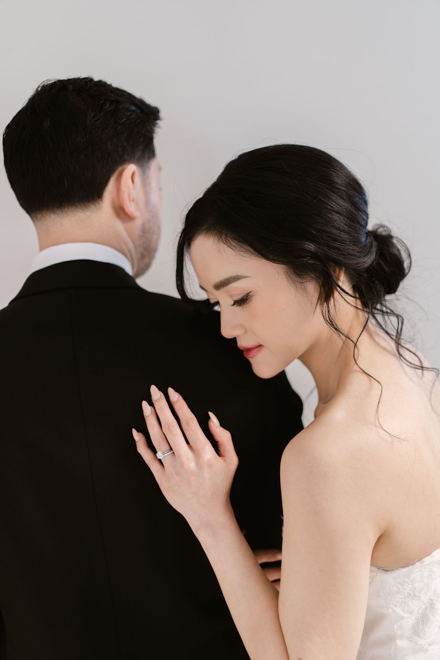 Bride in strapless lace gown rests hand on grooms black tuxedo shoulder, showcasing elegant engagement ring.