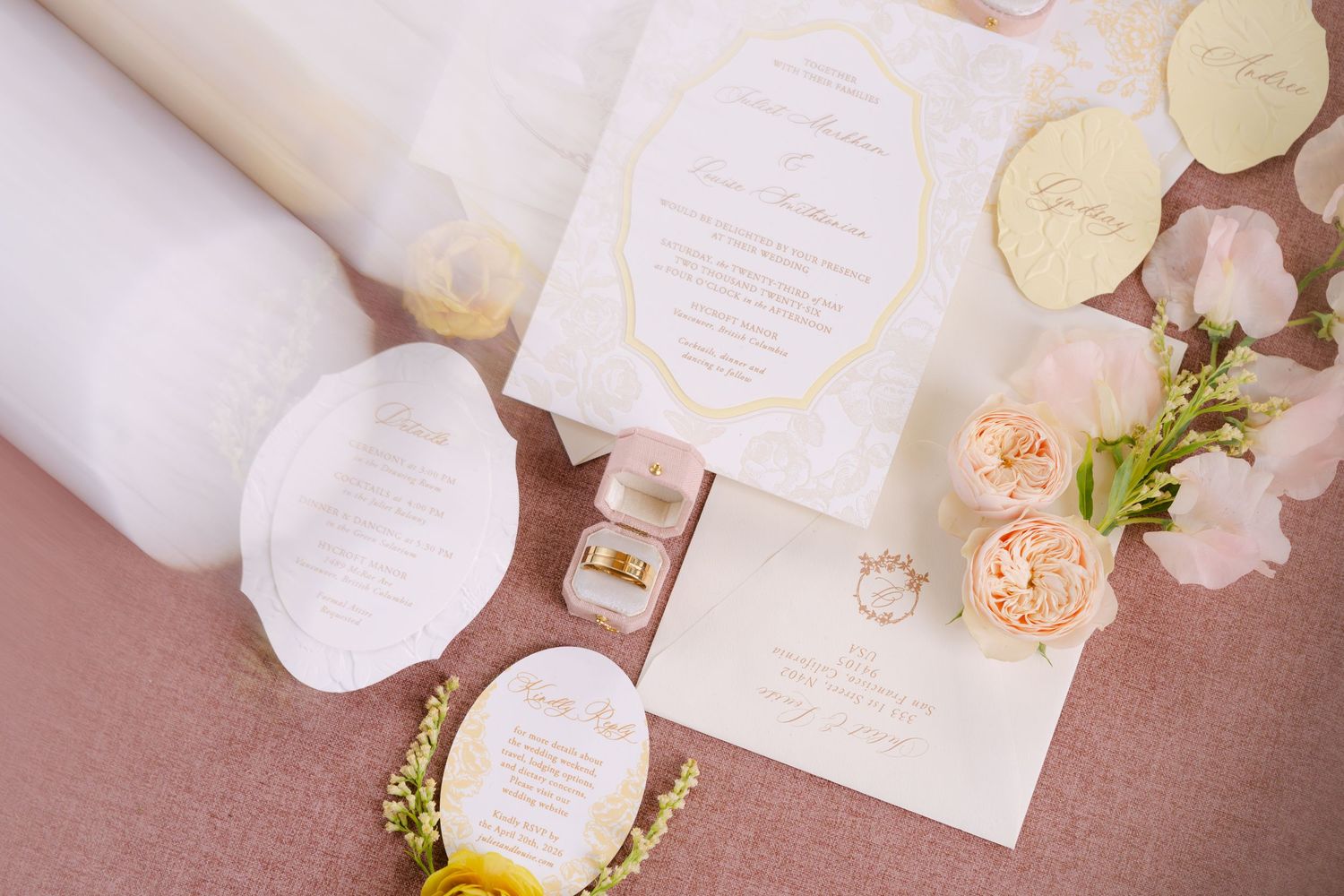 Wedding stationery flat lay featuring gold-trimmed invitations, rings, and peach roses on a blush pink surface.