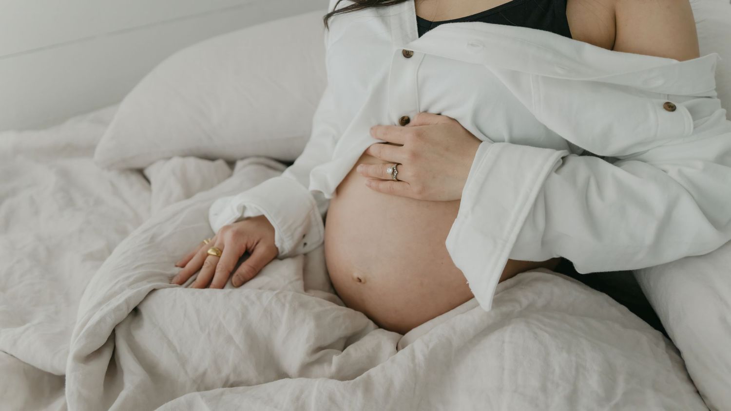 Close-up of pregnant belly with hands resting gently on white bedding.