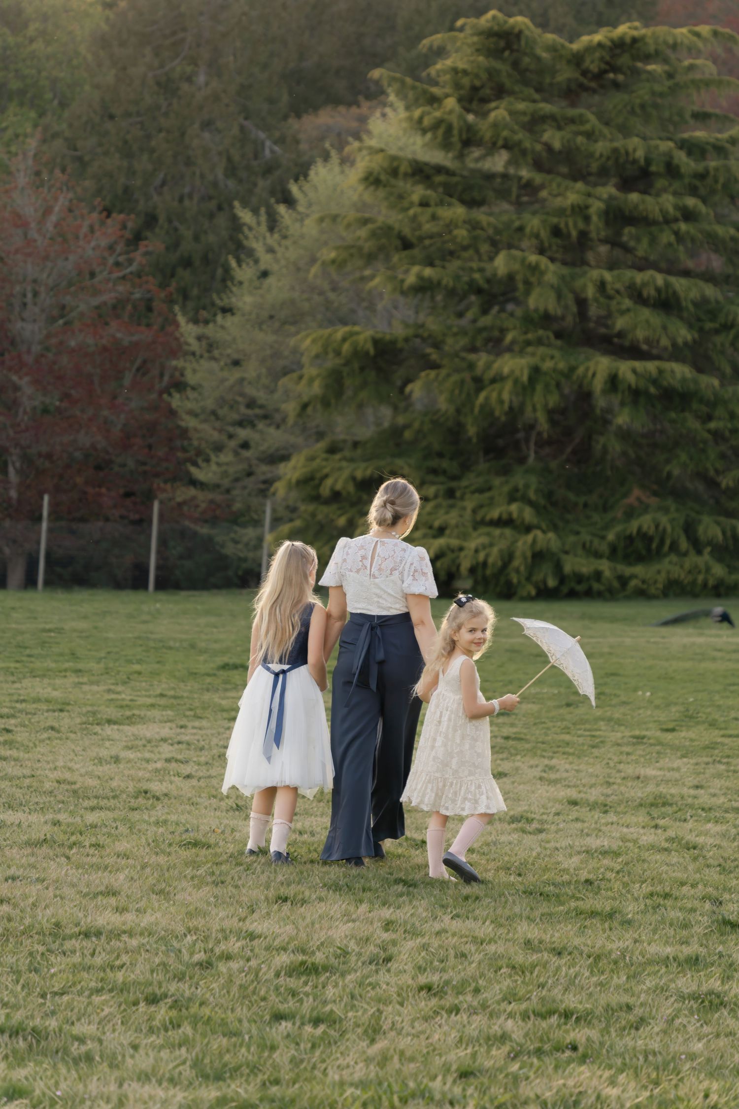 Three figures in light dresses stand in a grassy field with evergreen trees behind them.