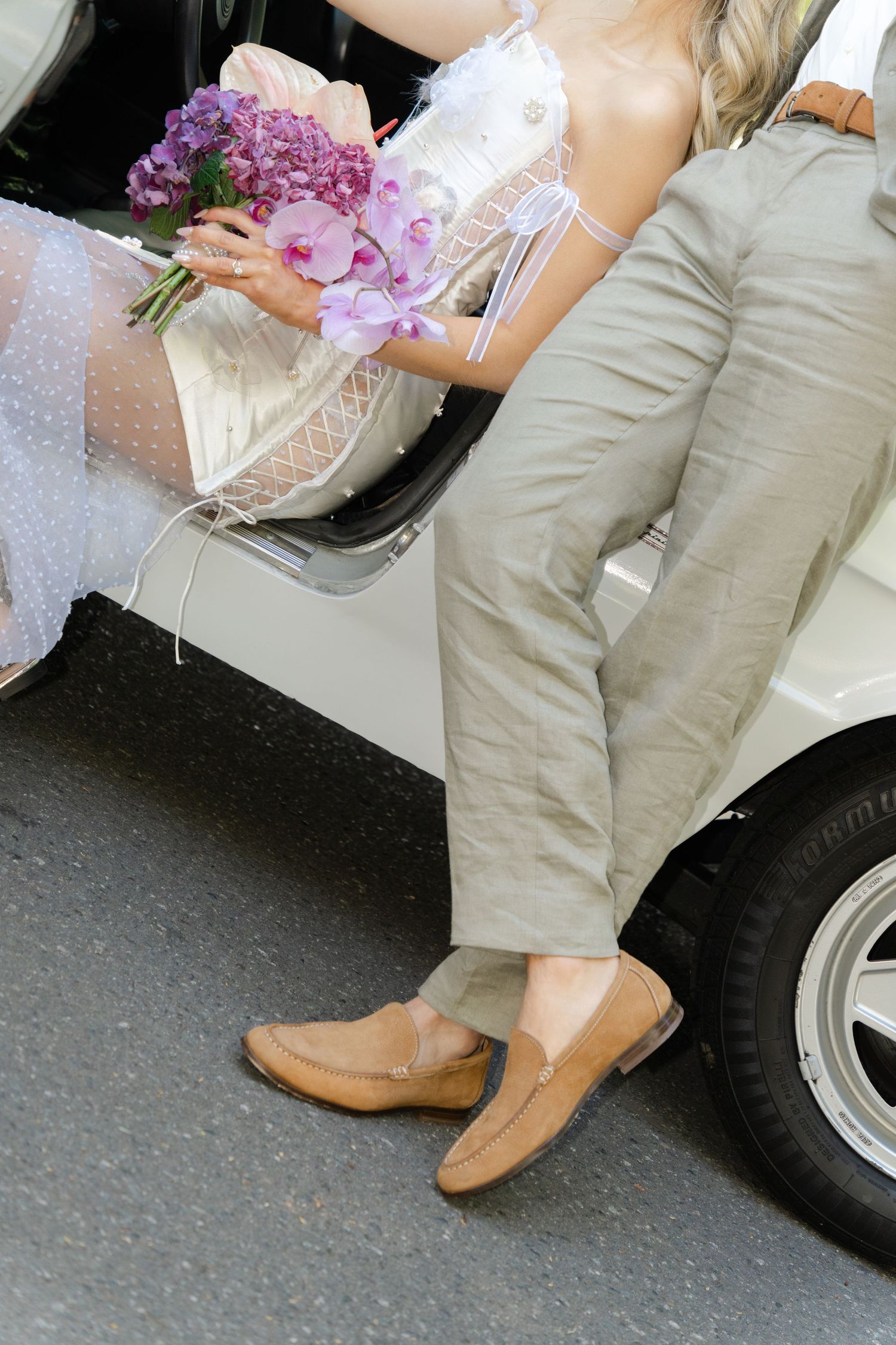Detail shot of beige loafers and purple bouquet next to car wheel.