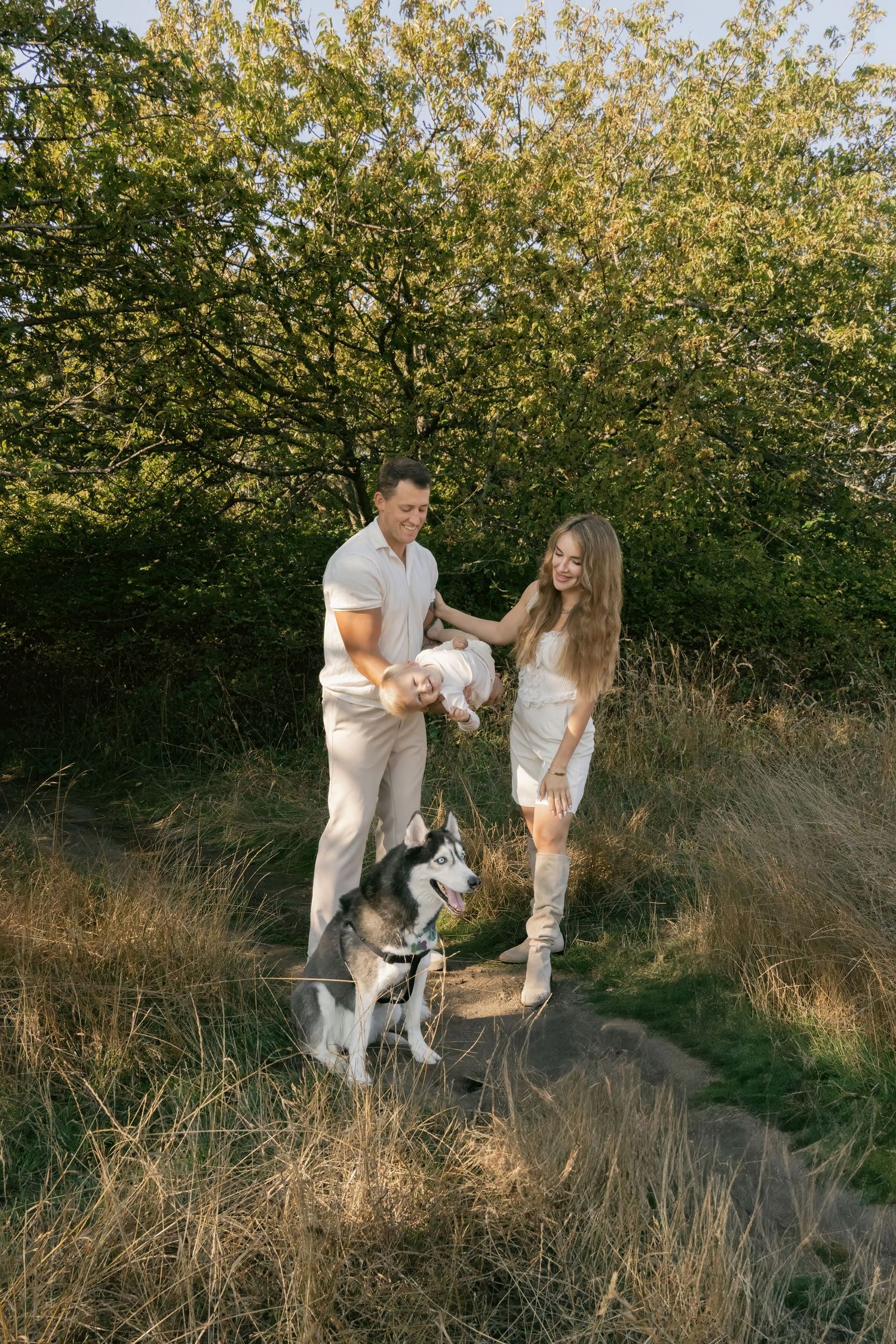 A couple in white outfits dances together in a golden meadow at sunset with their dog nearby.
