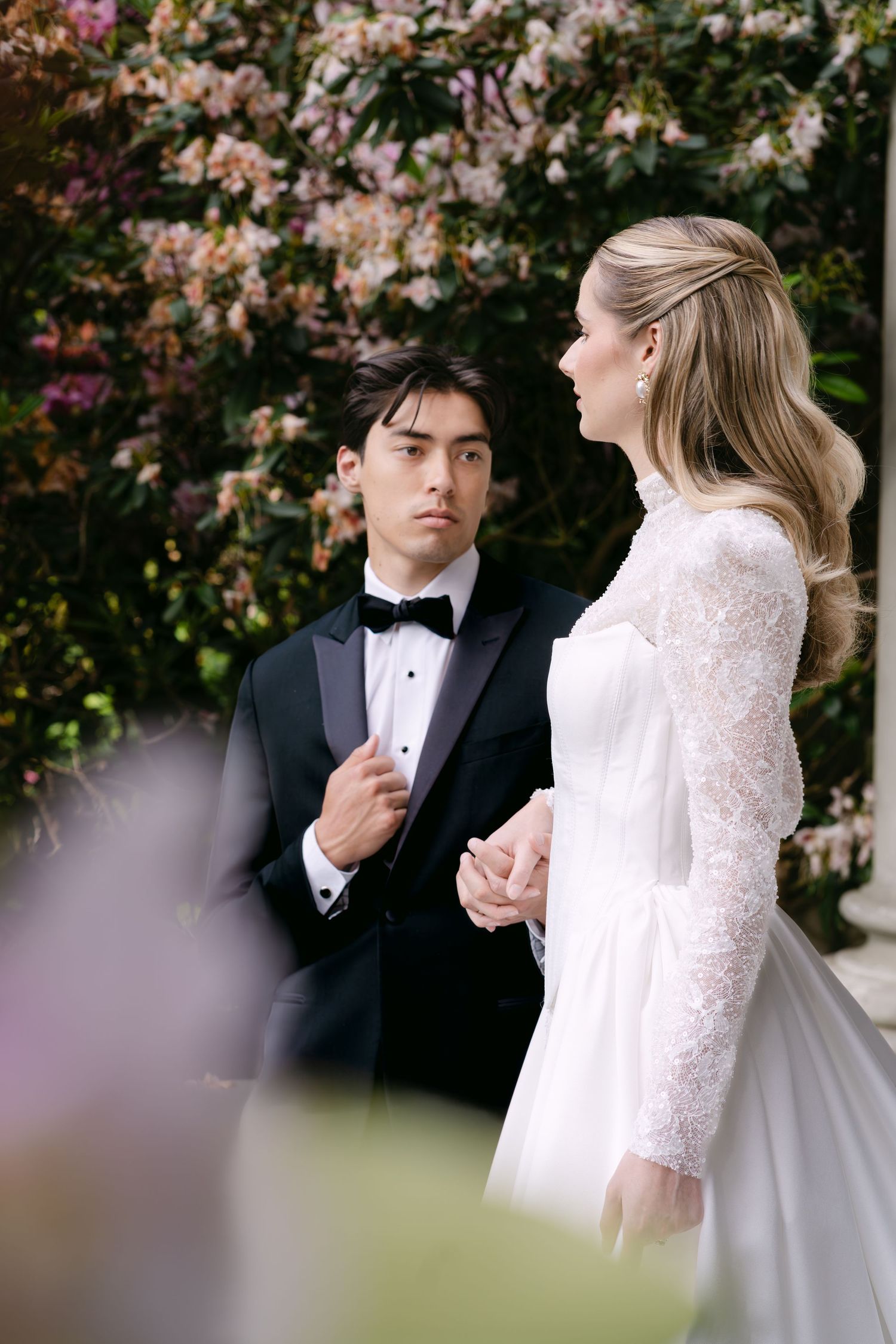 A couple in wedding attire holds hands among blooming flowers, the groom in a tuxedo and bride in a lace gown. In Vancouver