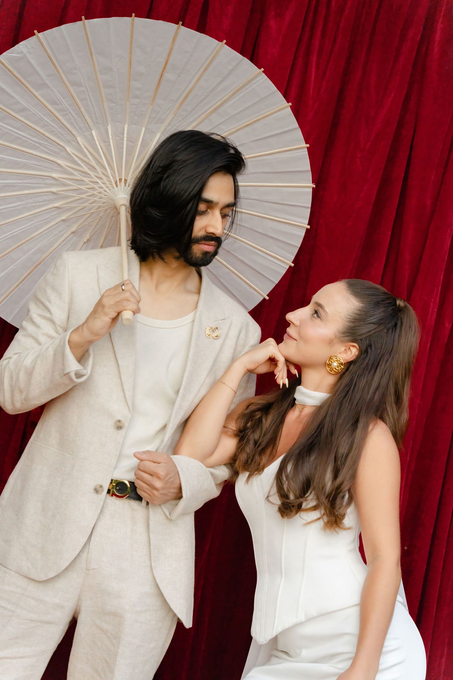 A stylish couple in white outfits poses against a red curtain backdrop, with one holding a decorative paper parasol.