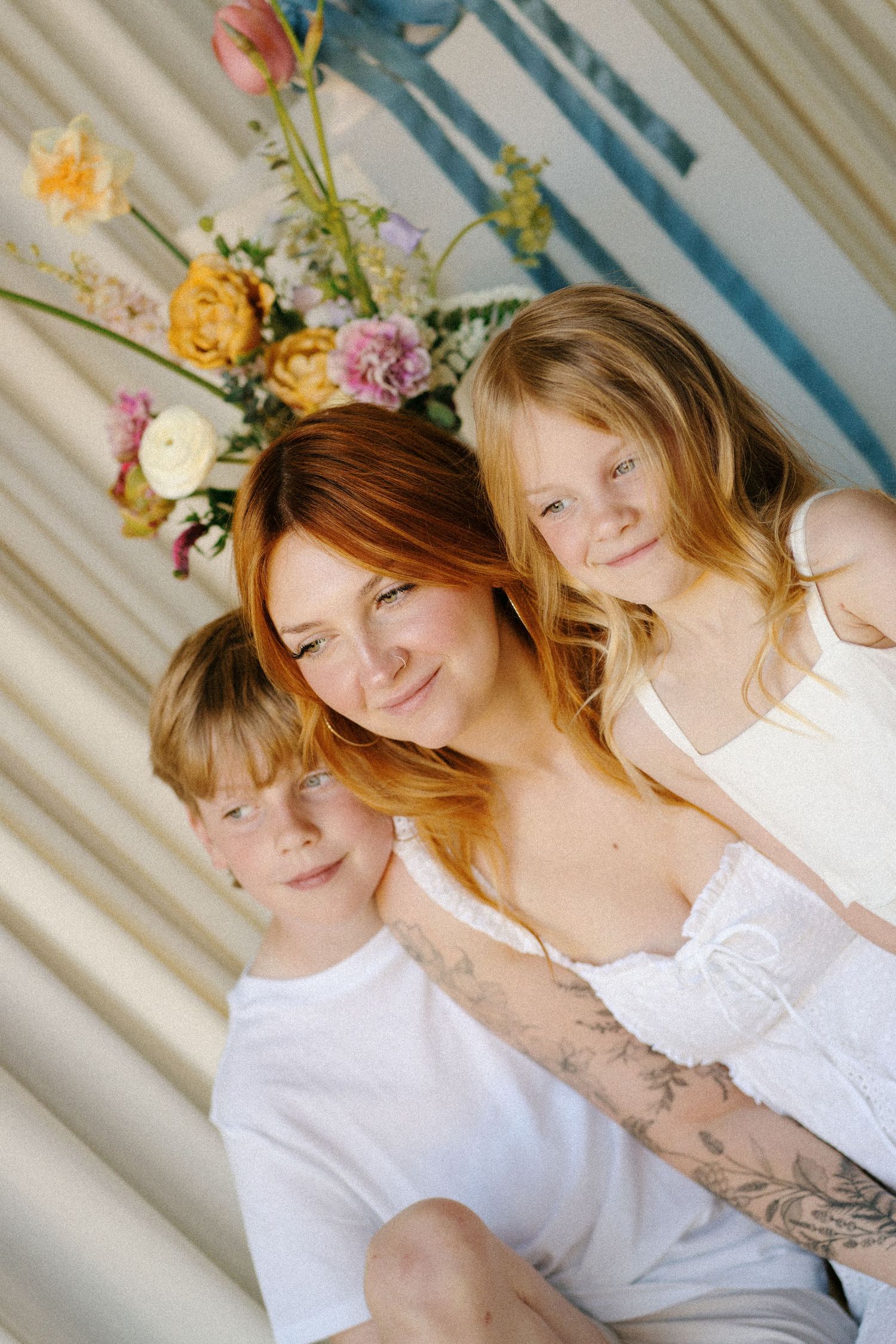 A redheaded woman with a tattoo smiles warmly while hugging two blonde children in front of colorful flowers.