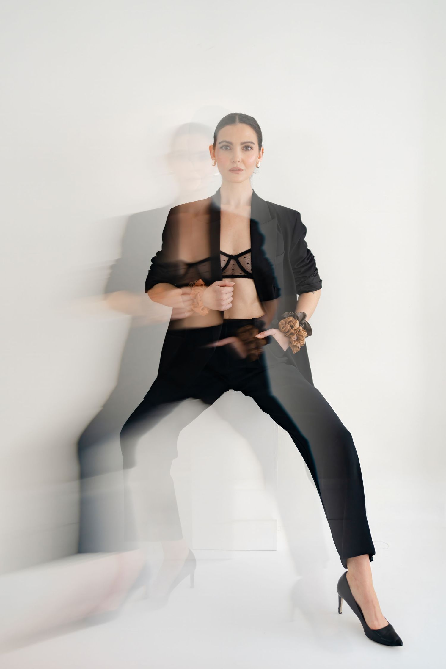 Woman in black blazer and heels poses confidently against white wall with artistic motion blur effect.
