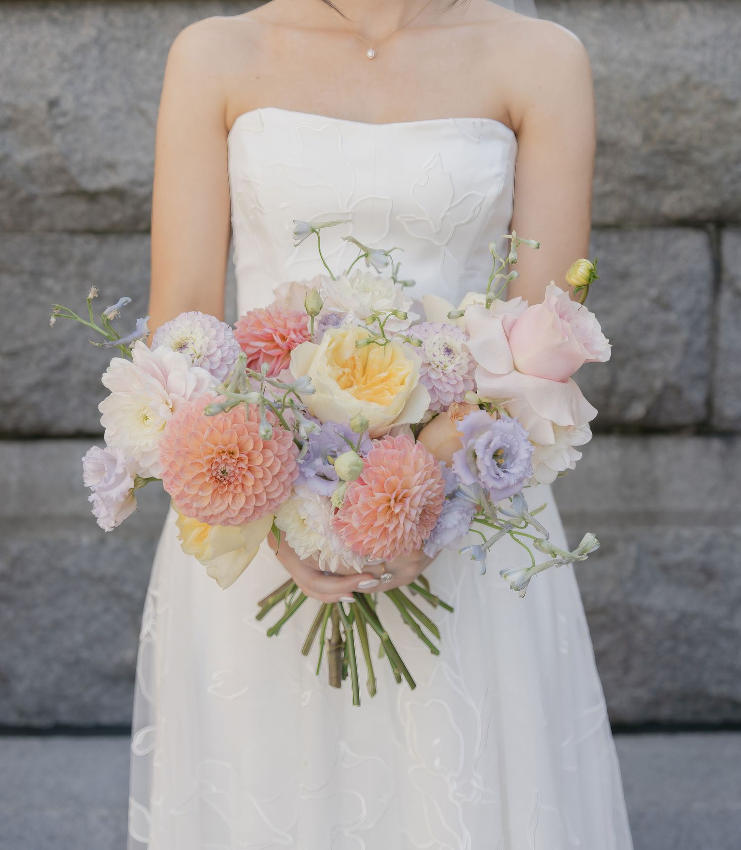 Pastel bridal bouquet featuring dahlias, roses and delicate spring flowers against stone wall.