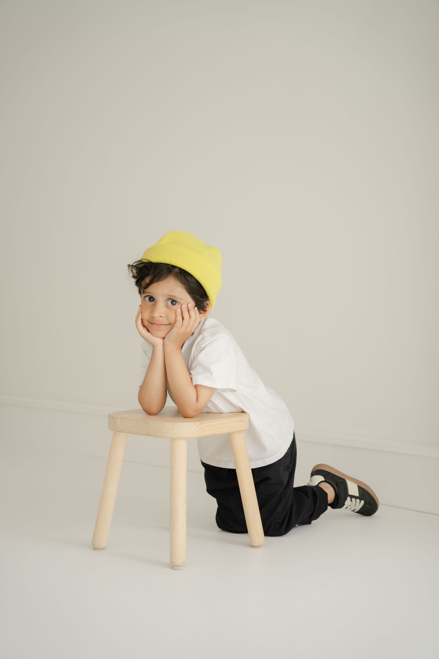 Playful kid in yellow beanie kneeling by wooden stool against neutral background.