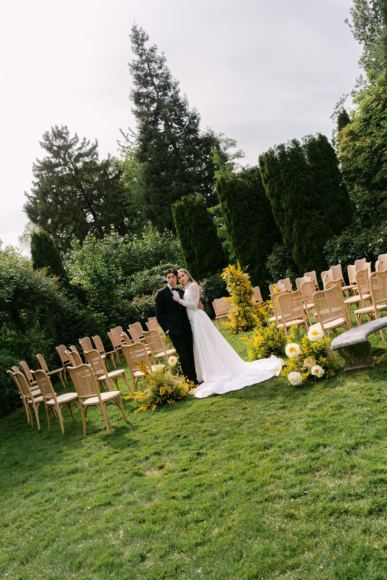Bride and groom embracing at outdoor garden wedding ceremony with wooden chairs and floral arrangements on green lawn.