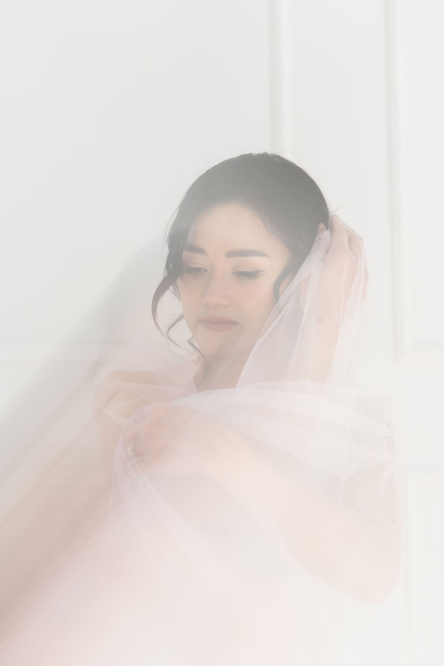 A bride wrapped in a soft white veil, gazing downward in a dreamy, ethereal bridal portrait.