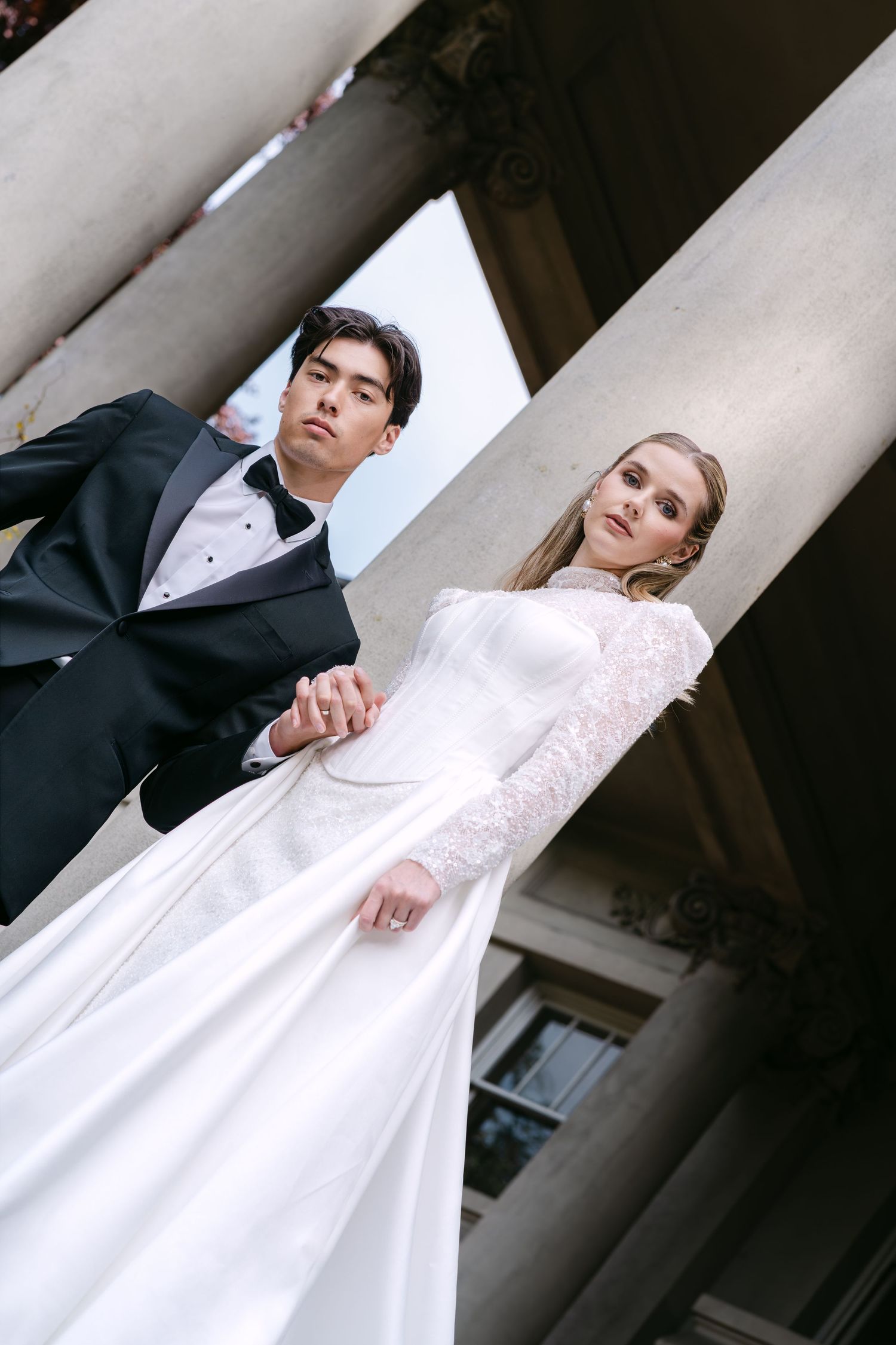 Elegant interracial wedding couple holding hands among classical columns, groom in tuxedo, bride in white lace gown.