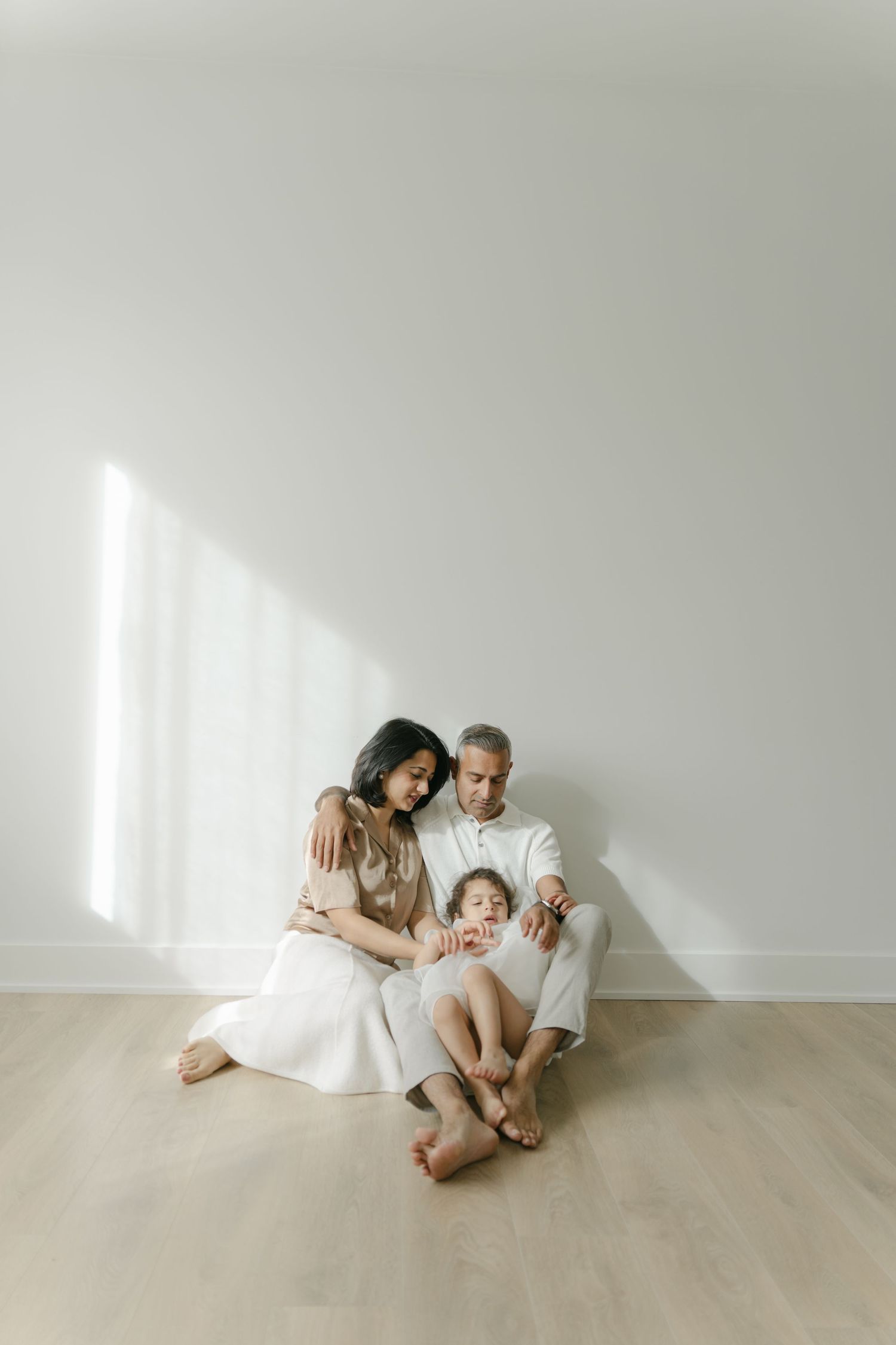 Family sitting together on wooden floor in minimalist white room with sunlight.