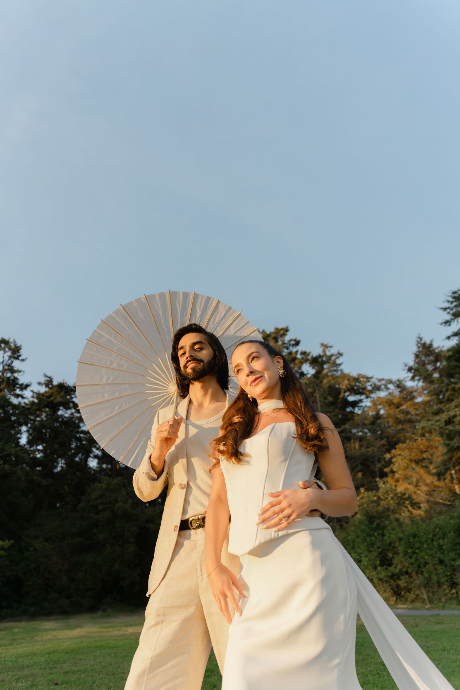 Two people in white outfits hold parasols while posing together outdoors during golden hour.