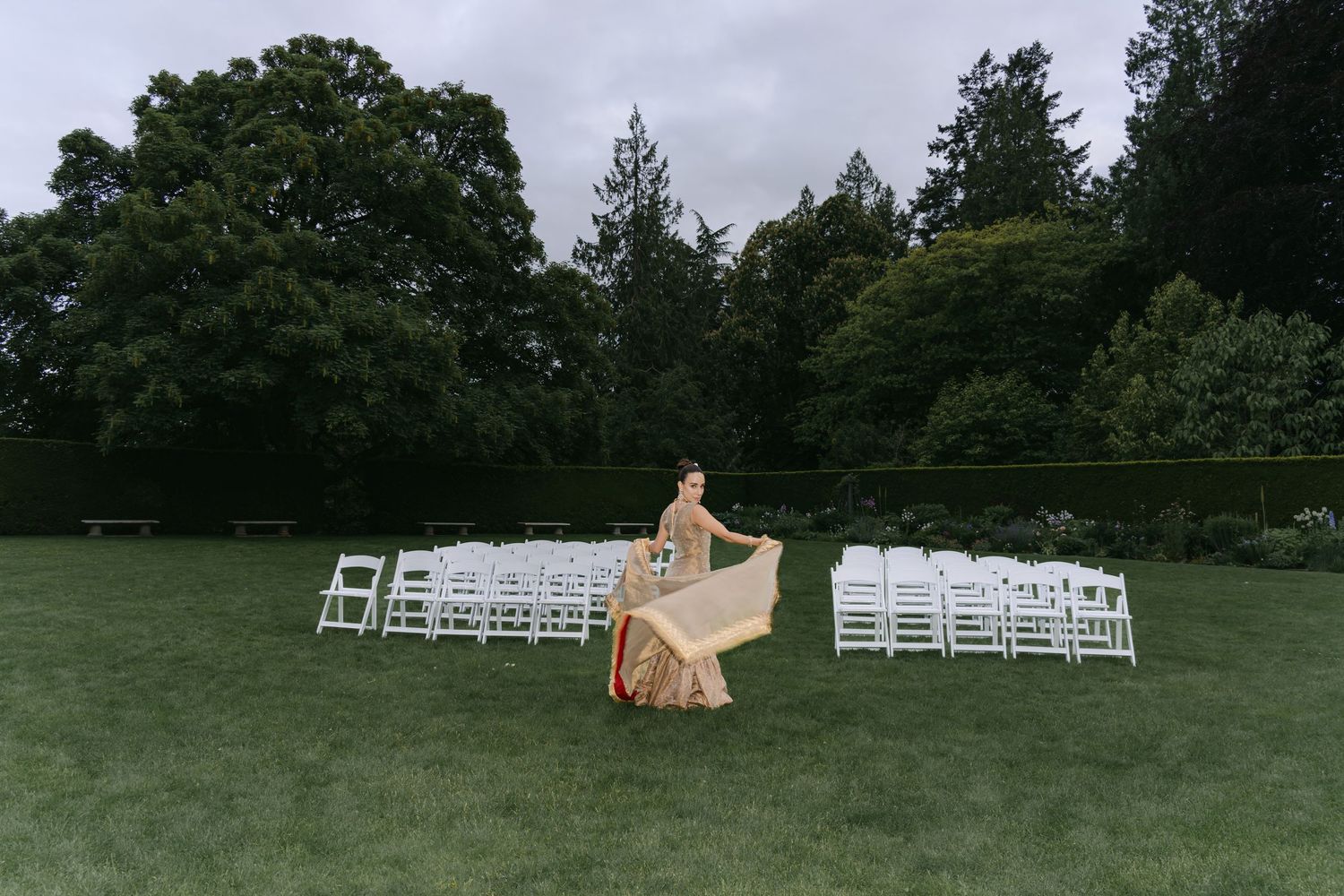 A bride twirls her gold gown on a lush green lawn with white chairs arranged for an outdoor wedding ceremony at Hatley Castle Victoria BC