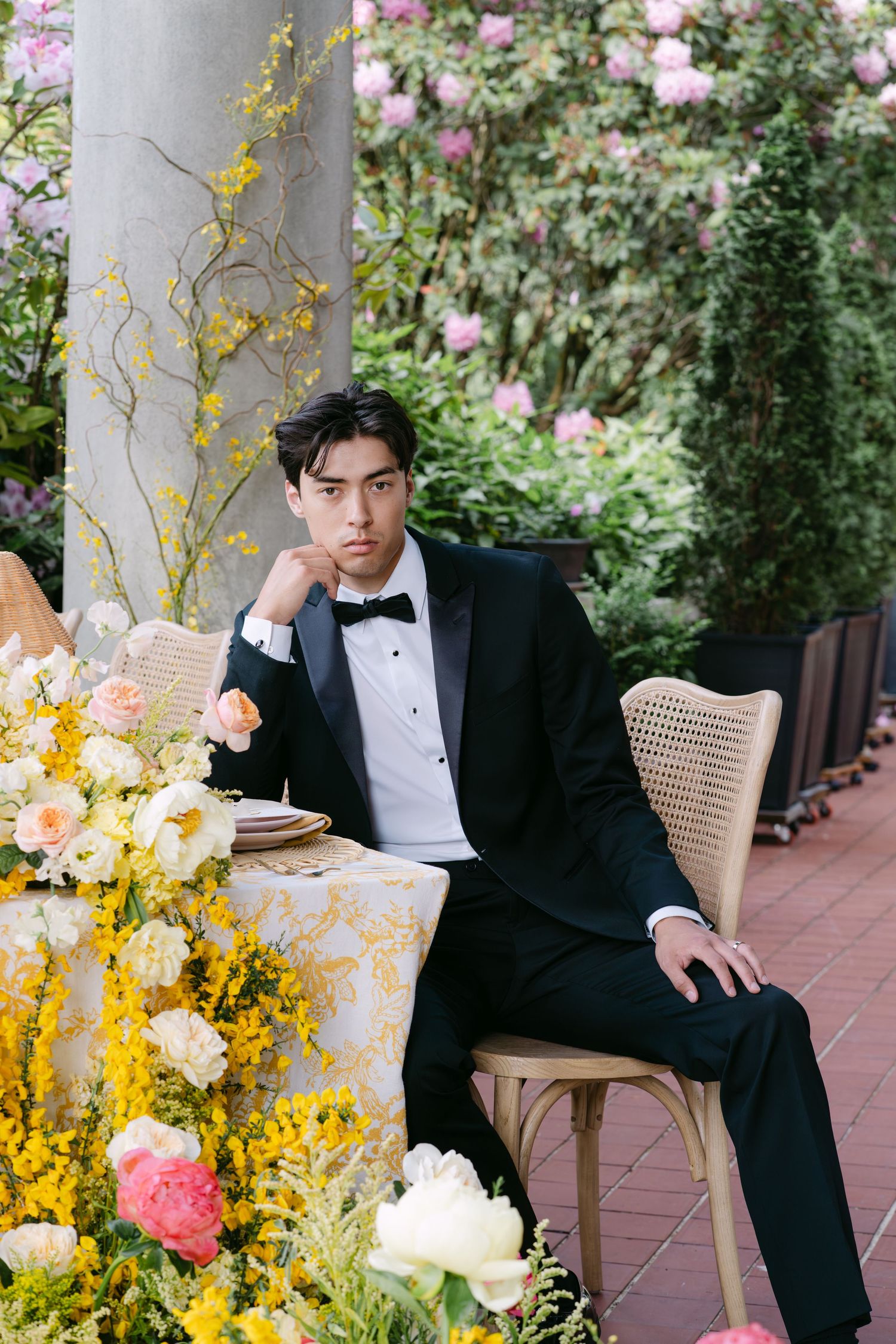 Groom in black tuxedo sits pensively at floral wedding reception table surrounded by yellow and pink blooms.