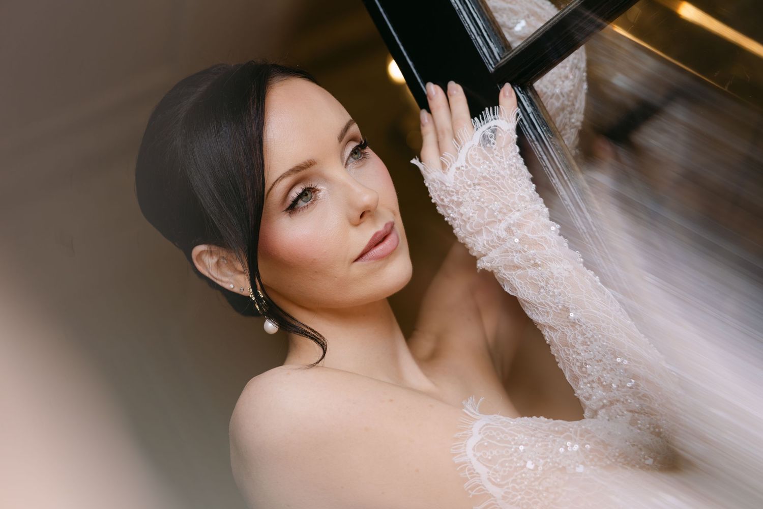 Beautiful bride with soft makeup and veil posing gracefully near a mirror on her wedding day.