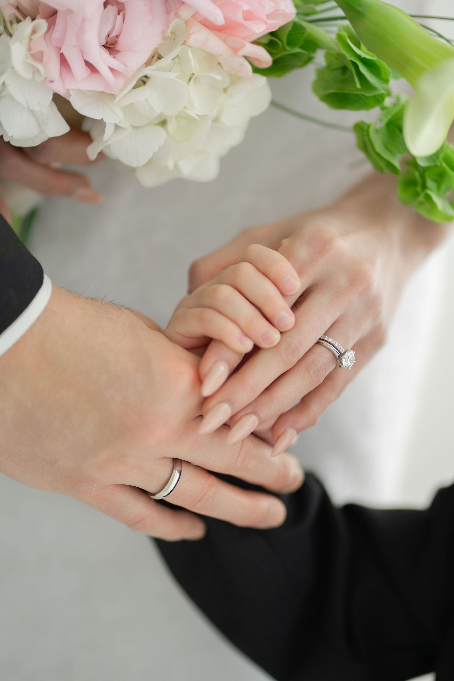 Newlywed couple holding hands with wedding rings, bride holding pink and white floral bouquet.