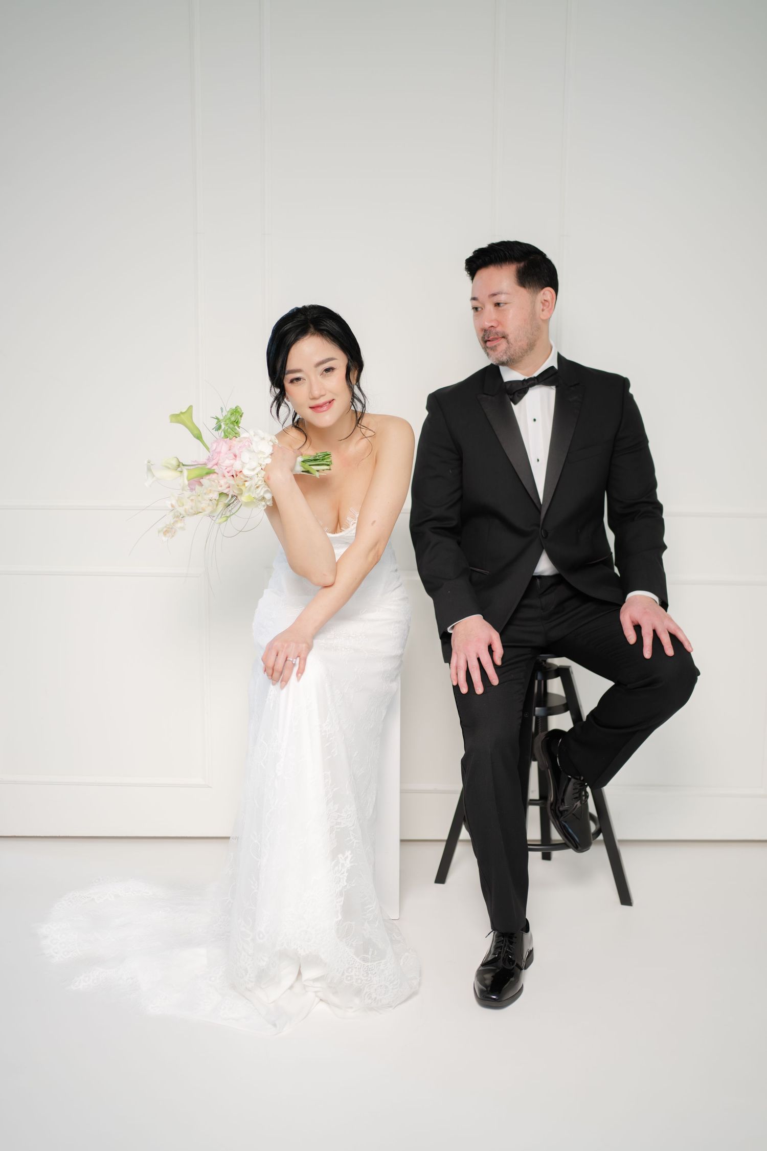 Bride in white gown holding pink bouquet poses with groom in black tuxedo on stool against white background.
