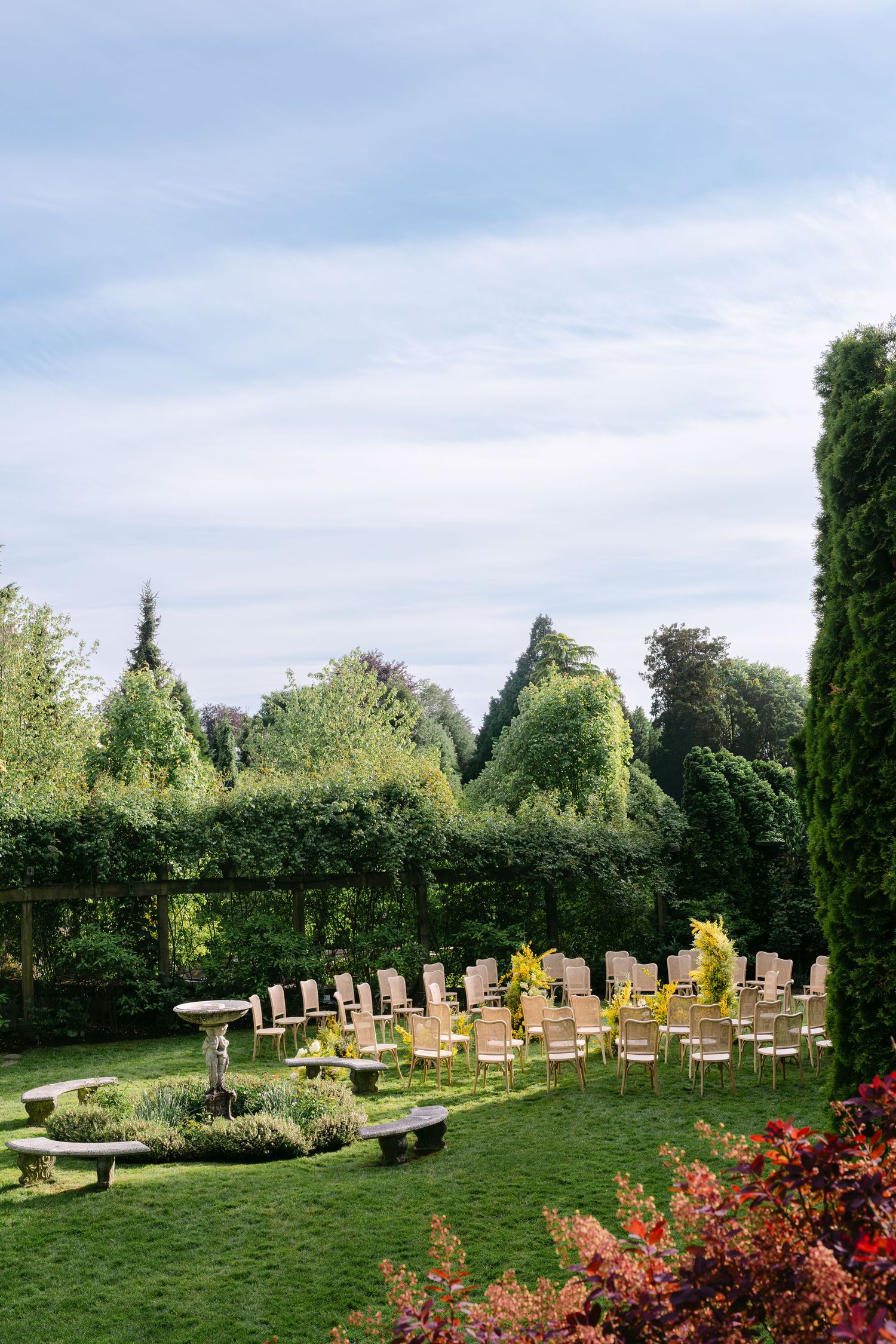 Elegant outdoor wedding ceremony setup with wooden chairs arranged on lush green lawn surrounded by manicured hedges and trees.