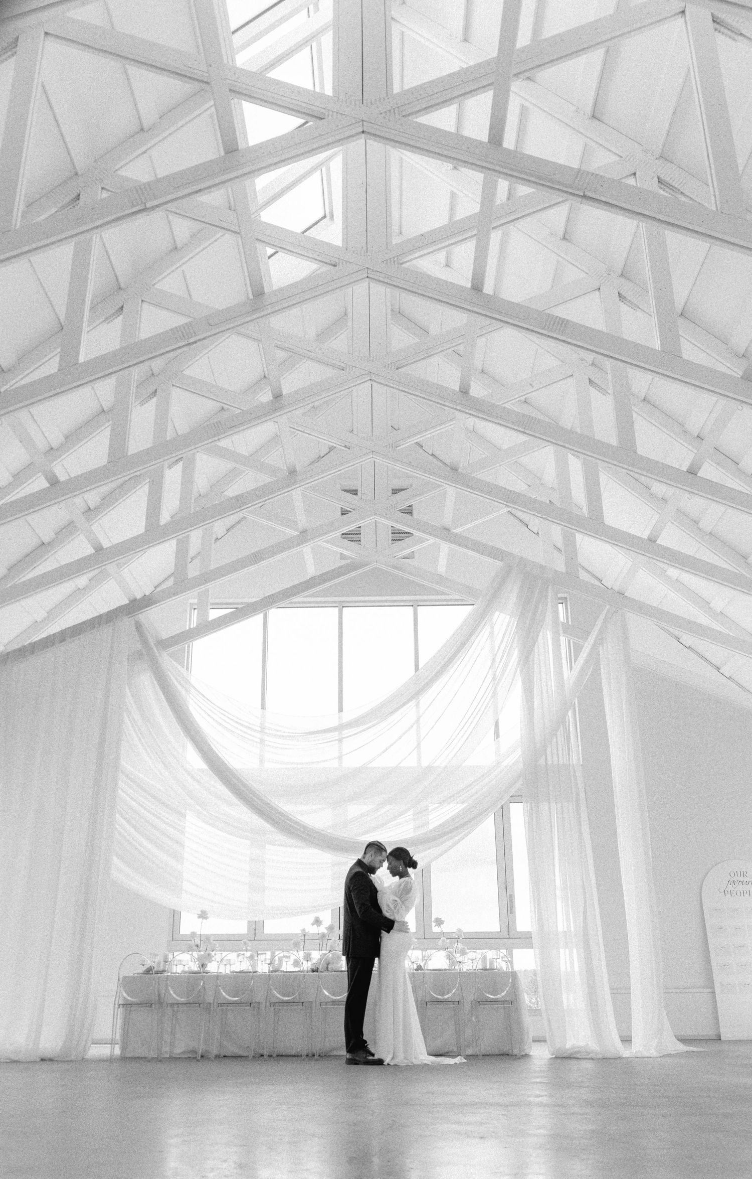 Black and white wedding photo of bride and groom embracing under draped fabric in elegant vaulted venue at Sea Cider Victoria BC.
