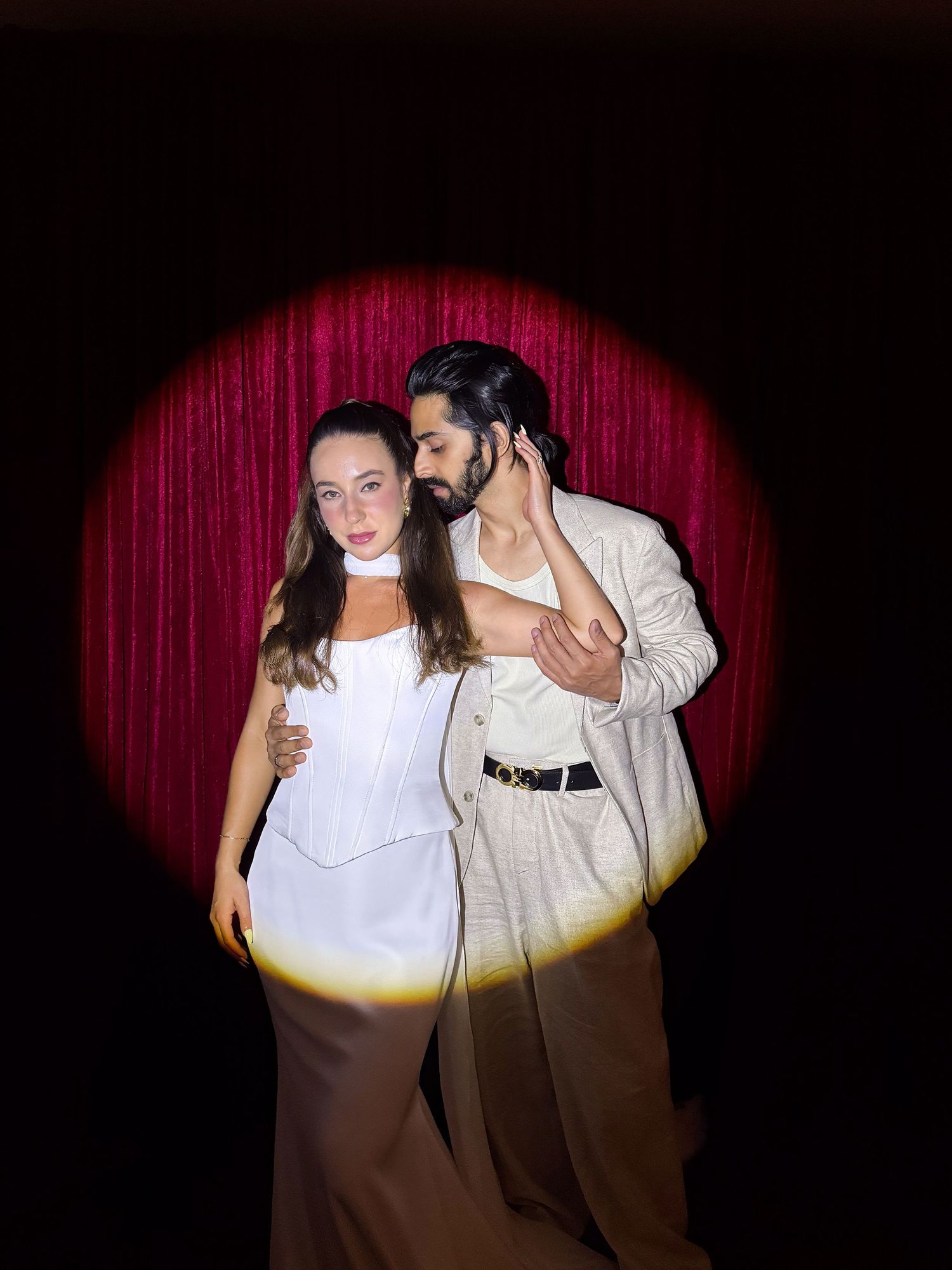 Two performers in brown and white outfits dance together under a spotlight against a dark red curtain backdrop.