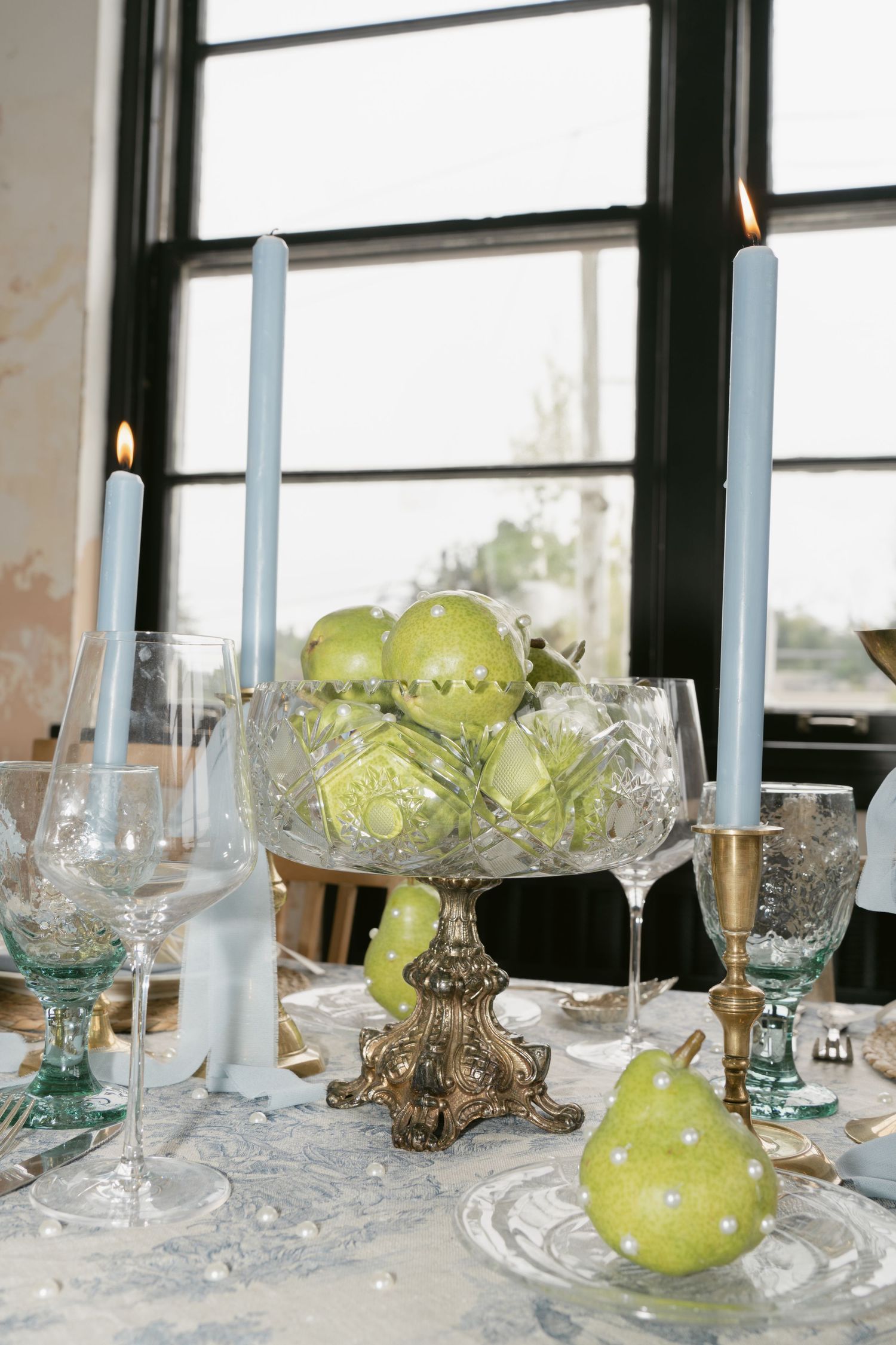 Crystal glassware and green decor arrangements on a table by sunlit windows.