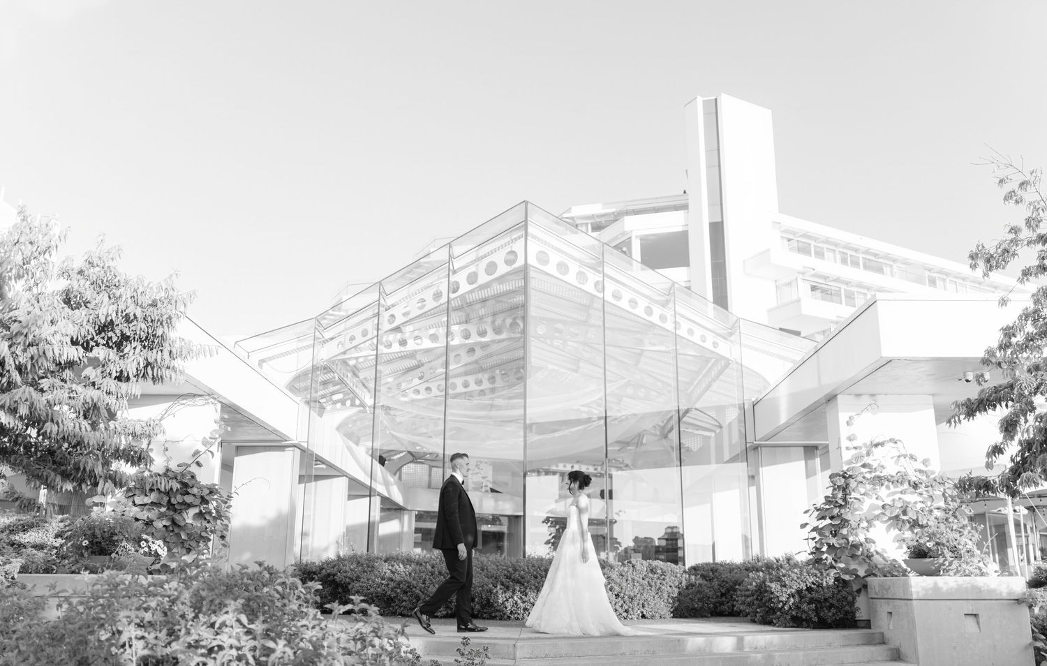 Bride and groom pose outside a modern glass wedding venue surrounded by lush greenery in a black and white photo.